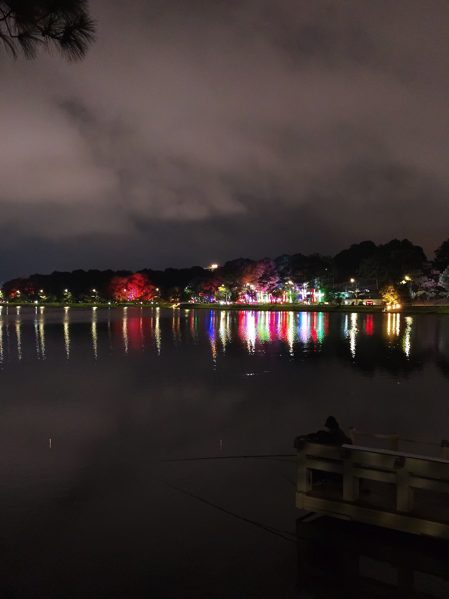 The image depicts a nighttime scene at Xuan Huong Lake in Da Lat, Vietnam. The lake reflects colorful lights from the buildings and trees lining its shore, creating vertical bands of red, blue, and yellow in the water. Overcast skies fill the upper portion of the frame. A wooden dock extends into the lake in the lower right. A person sits on the dock, their back to the camera, seemingly fishing as a fishing line stretches into the water. The foreground is dark, emphasizing the reflective qualities of the lake.