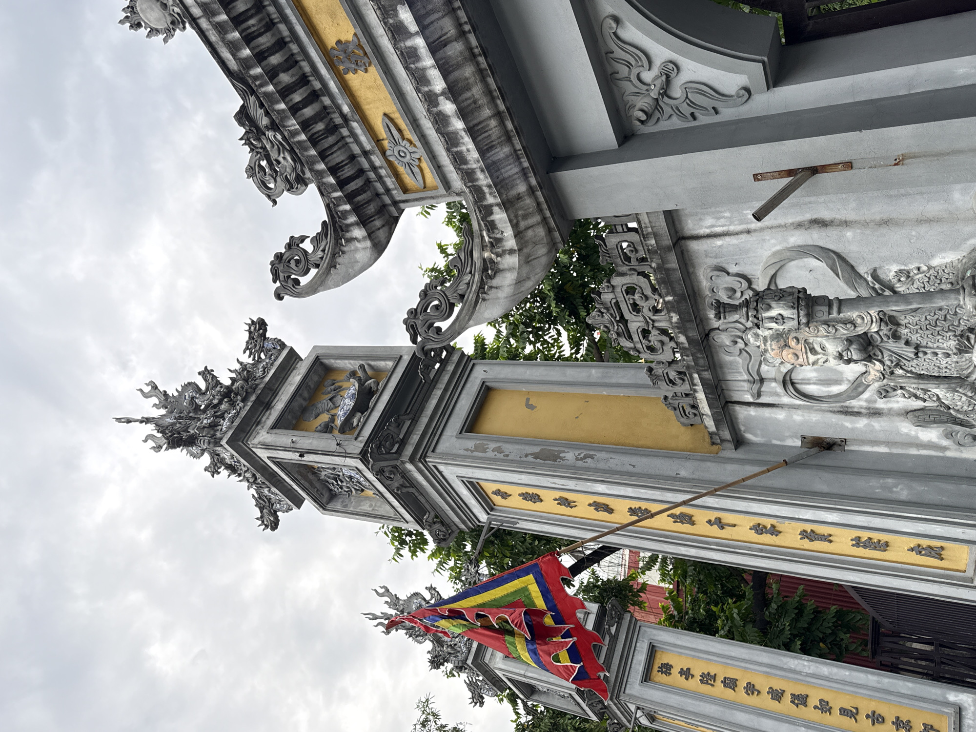 The image shows an ornate, gray stone and yellow-painted archway, likely the entrance to a temple or other religious site in Hà Nội, Vietnam. The archway has intricate carvings of dragons and other mythical creatures, along with classical Chinese characters on the yellow panels. A colorful, multi-paneled flag in shades of red, blue, green, and yellow hangs from a pole attached to the archway. The sky is overcast. The perspective is from below, looking upwards at an angle. Green trees and hints of other buildings are visible in the background through the arch.