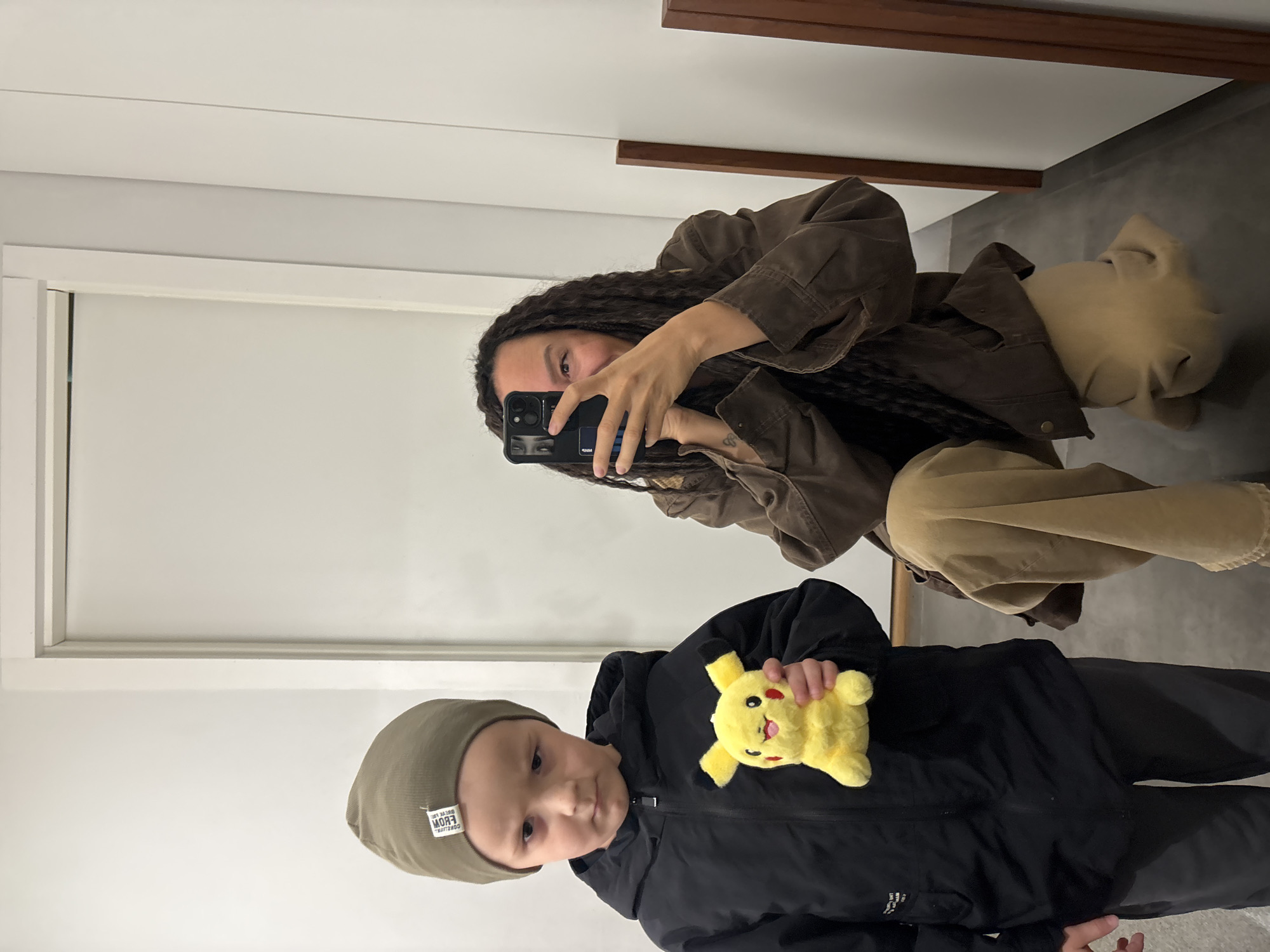 This mirror selfie, taken in Chaikovsky, Russia, captures an adult and a young child indoors. The adult, visible in the upper right of the frame through the mirror's reflection, is taking the photograph. She holds a black smartphone, which covers most of her face, with only one eye and part of her mouth visible, suggesting a neutral or slight smile. She has long, dark, braided hair and is dressed in a brown, heavy canvas or denim jacket over a dark top, paired with light tan or khaki-colored pants. A small, faint tattoo is discernible on her right wrist.
In the foreground, positioned lower and to the left, is a young male child. He is holding a bright yellow Pikachu plush toy, which has a wide, smiling face. The child is wearing a dark, possibly black, puffer jacket and an olive-green, rib-knit beanie. A white fabric tag on the beanie features mirrored black text. When unmirrored, the text reads "FROM / COFFEE LIFE / NEW YORK". The child looks directly towards the camera with a calm, neutral expression.
The indoor setting features pristine white walls, with a white doorframe visible on the left side. The floor appears to be a dark gray or concrete, and a decorative brown wooden beam or trim runs horizontally across the top of the reflected wall. The lighting is bright and even, consistent with an indoor environment. The scene conveys a casual, everyday moment, possibly captured in a home or a public space with a large mirror.