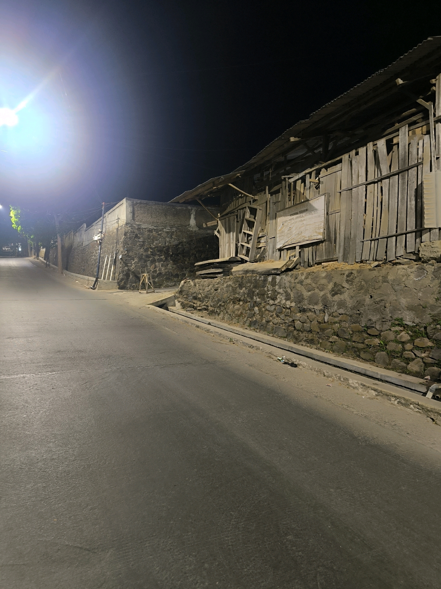 The image depicts a street scene at night in Bandung City, Indonesia, characterized by an overhead light source creating a bright glare on the left side of the frame. The street is paved and curves to the right, with a slight incline. On the right side of the street, a tiered structure is visible, built into a hillside. The lower level is a stone retaining wall, topped by a wooden structure with a corrugated metal roof. A sign with text is affixed to the wooden wall. A small drainage channel runs along the edge of the road. The overall atmosphere is dimly lit, with shadows cast by the streetlights and the structures.