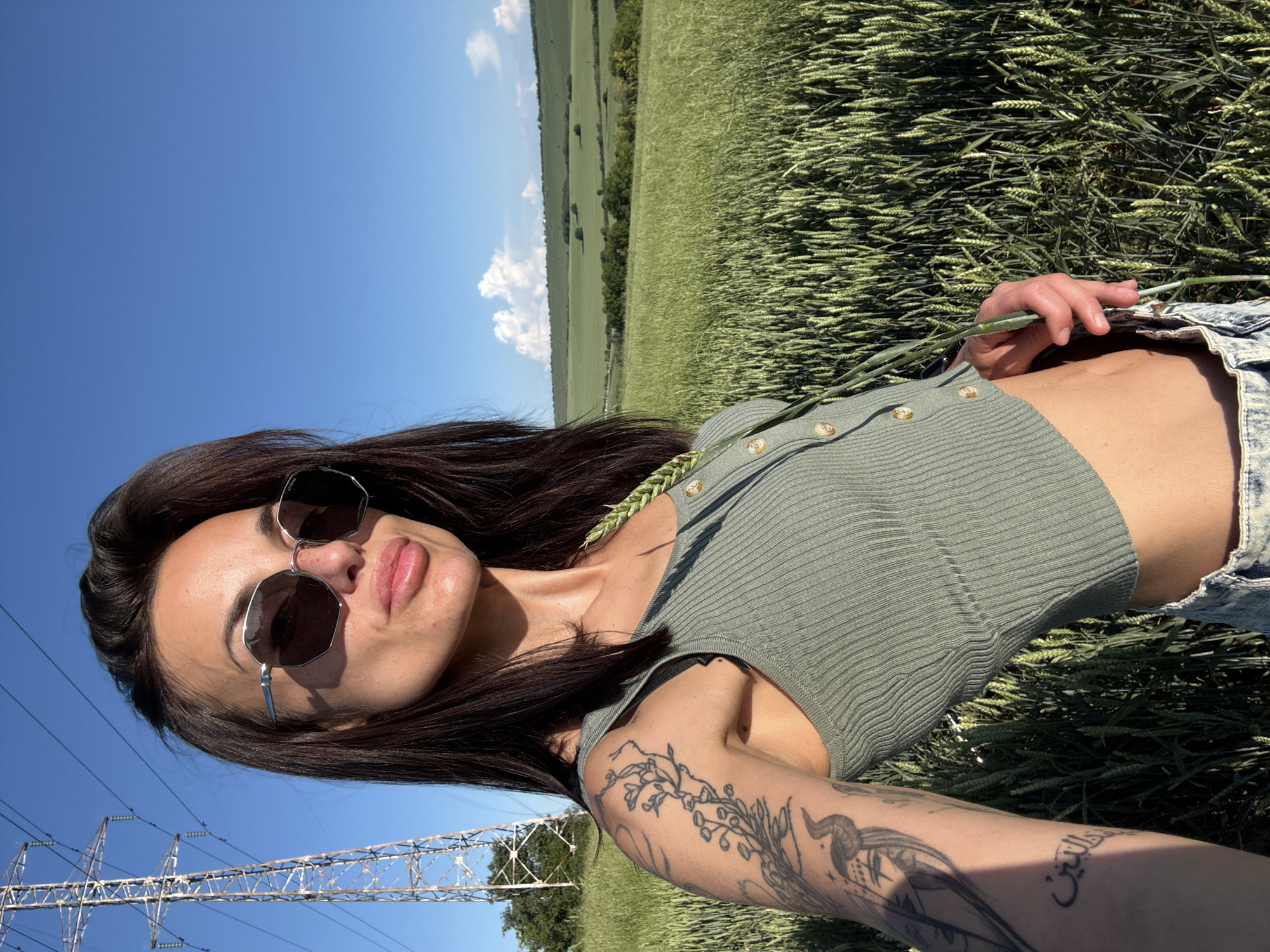 A woman with dark hair and sunglasses poses in a field. She wears a green ribbed crop top and denim shorts. Her left arm features tattoos. She holds a stalk of wheat. The background shows a field of wheat under a blue sky with clouds. In the distance, a tree line is visible. In the lower left, there is a power line tower. This field could be located in or around Pleven, Bulgaria, which is known for its agricultural land.