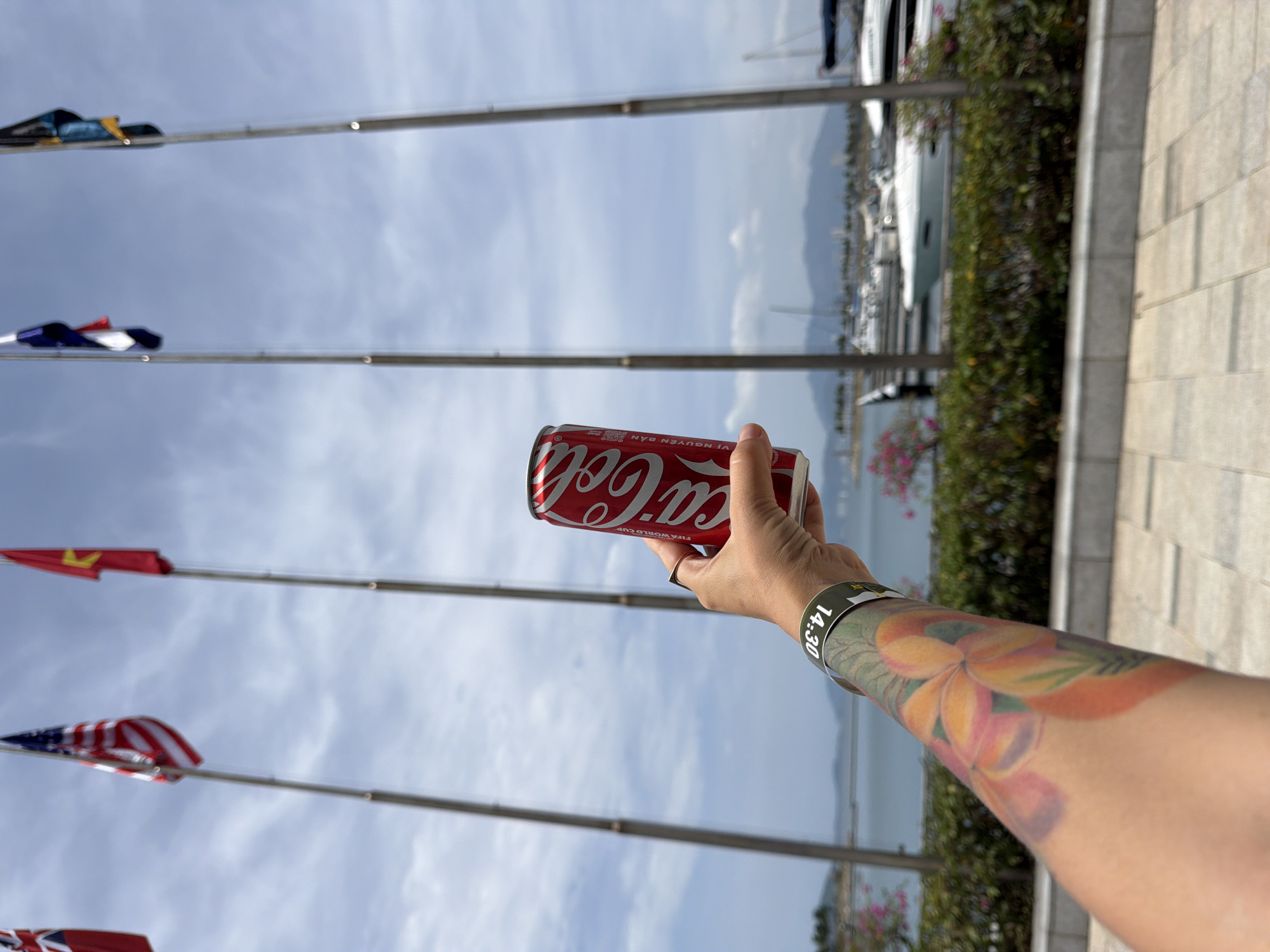 A person's tattooed arm holds a red Coca-Cola can in the foreground, with a wristband reading "14:30" visible. Behind the arm, several national flags fly on tall poles against a cloudy sky. In the middle ground, a body of water with numerous yachts and boats is visible, with a treeline and distant mountains forming the backdrop. The scene is set outdoors, likely in a harbor area of Nha Trang, Vietnam.