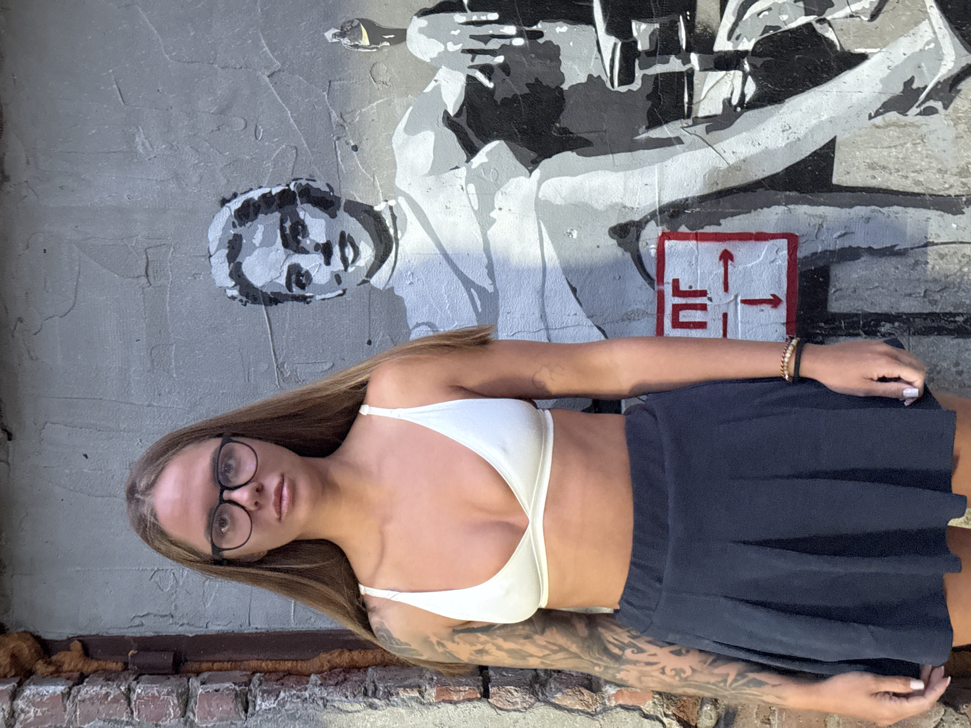 A young woman stands in front of a mural on a wall in Nizhny Novgorod, Russia. The mural depicts a man aiming a rifle, next to a red and white sign with arrows pointing in multiple directions. The woman wears a white top and a dark skirt. She has long brown hair and glasses. A brick baseboard runs along the bottom of the wall. The image is taken outdoors, likely in daylight.