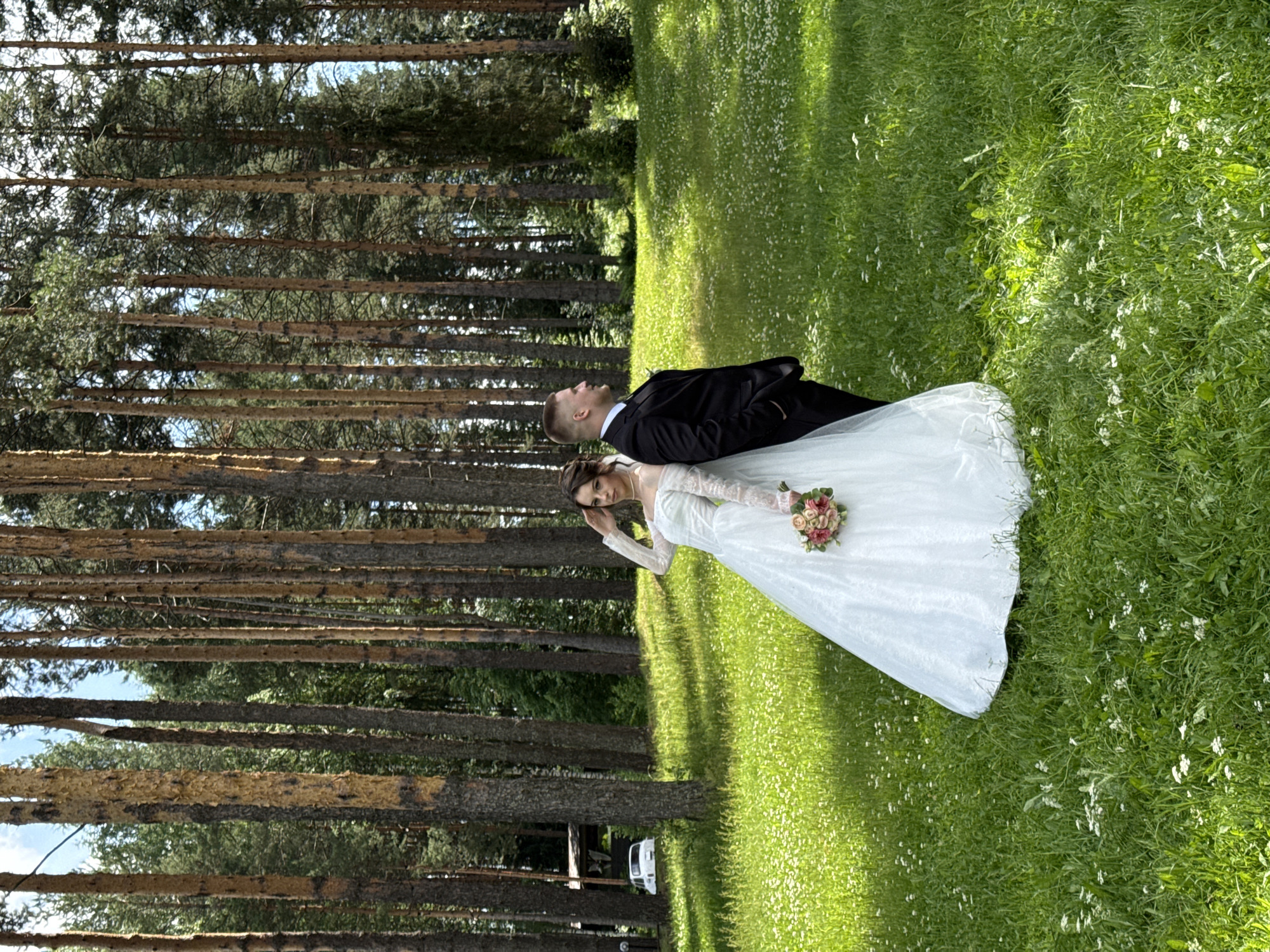 A wedding portrait of a bride and groom standing back to back in a sunlit forest clearing. The bride, wearing a white off-the-shoulder wedding dress with long lace sleeves and holding a bouquet of pink and cream roses, is positioned on the left side of the frame. The groom, in a black suit and white shirt, stands to her right. They are positioned in a grassy area dotted with small white wildflowers, with a dense forest of tall pine trees forming the background. The light filters through the trees, creating dappled shadows on the ground. In the background, a white car is visible parked near a small wooden structure. The sky is partly cloudy. This scene is located in the city of Votkinsk, Russia.