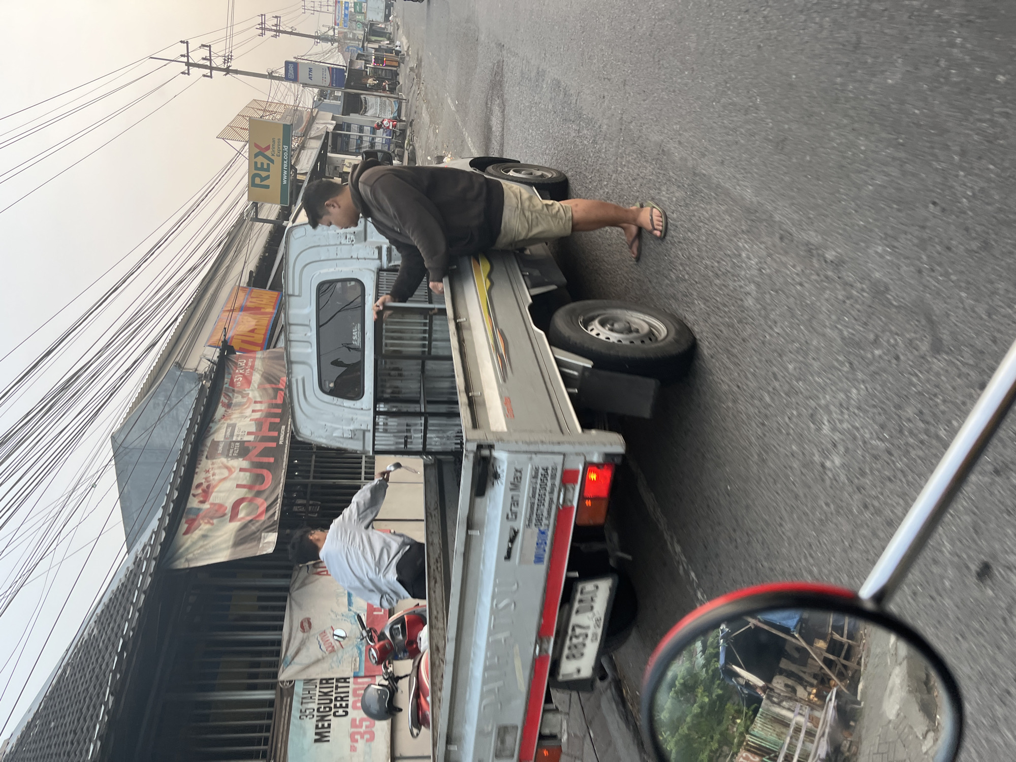 In Kludan, Indonesia, a man wearing a brown hoodie and khaki shorts stands beside the bed of a silver Daihatsu Gran Max pickup truck. He is holding a small silver object, possibly a tool or a piece of debris, and appears to be interacting with the truck's cargo area. Another person, wearing a light blue long-sleeved shirt, is leaning into the truck bed from the driver's side. The truck is parked on a paved road lined with buildings and overhead power lines. Banners and signs, including one for "REX Kiriman Express" and another with "DUNHILL", are visible on the buildings. The scene suggests a mundane street activity, possibly loading or unloading goods, in a commercial or residential area of the town.
