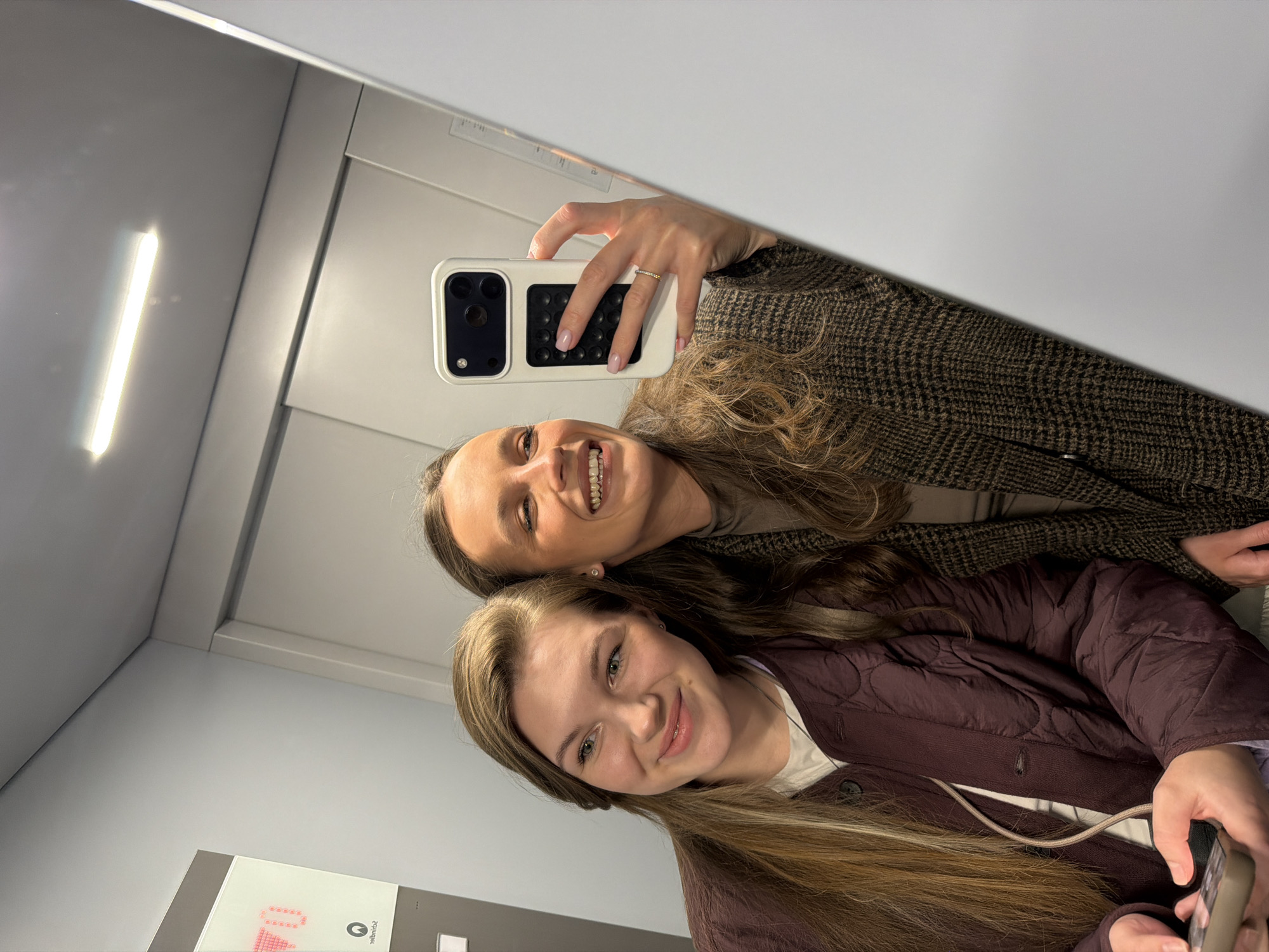 Two women are taking a selfie in an elevator. The woman in the foreground has long blonde hair and is wearing a burgundy quilted jacket. The woman in the background has curly brown hair and is wearing a brown herringbone jacket. The woman in the background is holding a white smartphone with a pop-socket on the back. The elevator is plain and white with a fluorescent light. A panel with illuminated numbers and a logo is visible on the elevator wall. There is no information available to suggest that the image was taken in the city of Vozdovac Urban Municipality, Serbia.