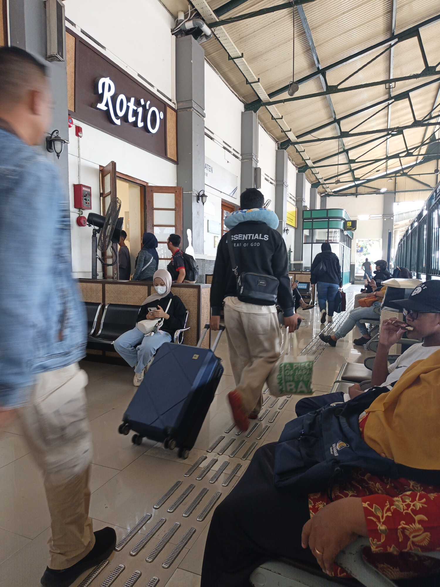 The image depicts a scene inside a train station in Klaten, Indonesia. In the foreground, a person is walking from left to right, with their back partially visible, wearing a blue denim jacket and light-colored pants. They are pulling a dark blue suitcase on wheels. To the right, several people are seated on benches, waiting. One individual, wearing a light-colored headscarf and a white face mask, is looking at their phone. Another person in the foreground, on the right side of the frame, is wearing a dark hat and appears to be eating.
In the background, there is a sign for "Roti'o" above a doorway, suggesting a food establishment within the station. The station has a covered roof supported by metal beams, and the floor is tiled. There is a fan mounted on a stand near the doorway. The overall atmosphere appears to be that of a busy public transportation hub during the daytime.