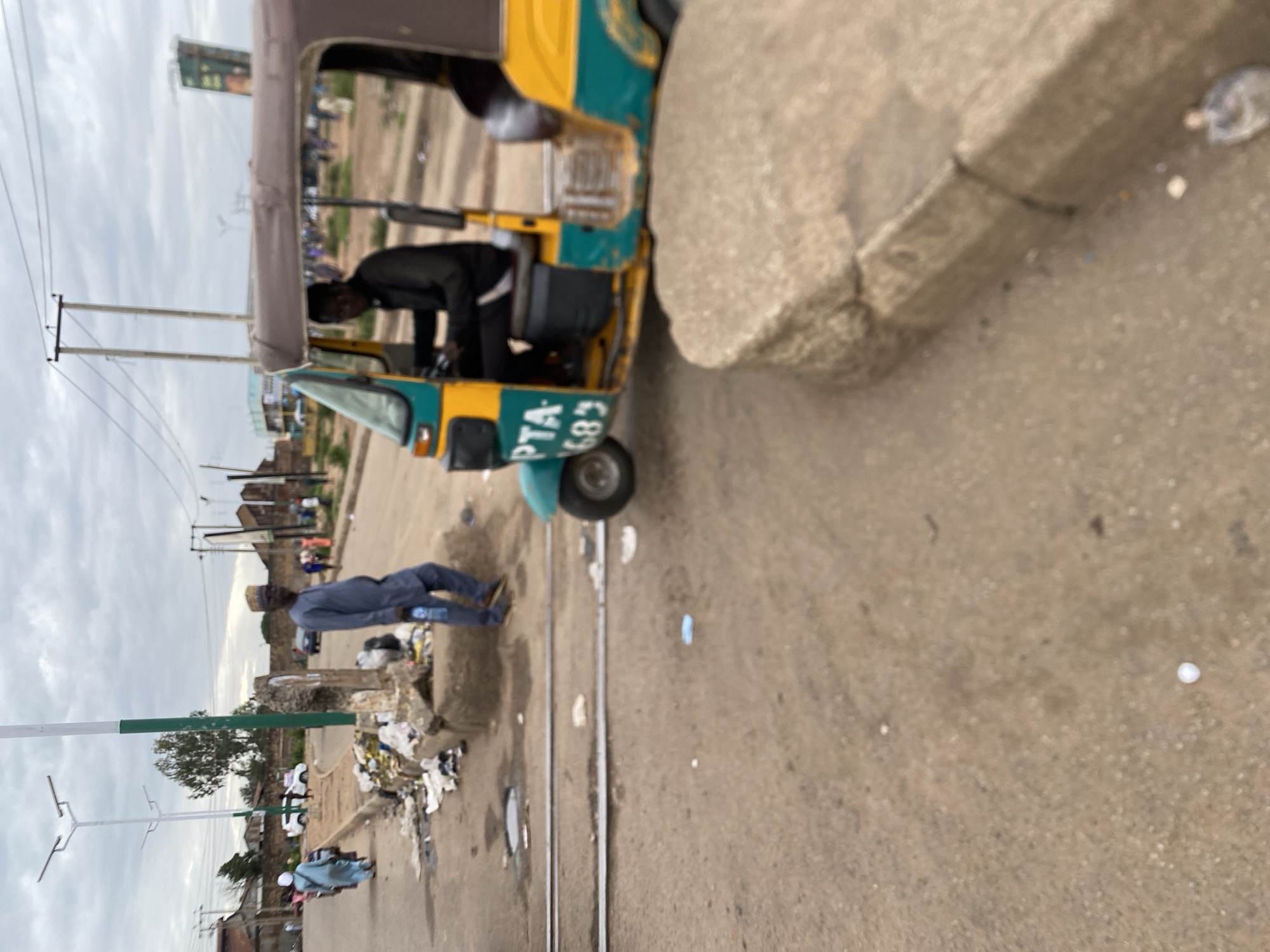 The image shows a street scene in Bukuru, Nigeria. The primary subjects are a yellow and green auto-rickshaw, likely a form of public transportation, and two men. One man, wearing a dark jacket, is seated inside the auto-rickshaw, seemingly in the driver's position. The other man, dressed in a blue shirt and pants, stands outside the vehicle, holding a clear plastic bottle. The auto-rickshaw has visible text on its side: "PTA 683".
The setting appears to be an urban or semi-urban area with buildings in the background. There is a concrete curb and a dusty, paved road, suggesting it is daytime. The sky is overcast, indicating a cloudy or possibly rainy weather condition. The ground near the curb is littered with trash, including plastic bags and wrappers, which is a notable detail of the scene. The overall impression is one of everyday life in a Nigerian town, with transportation and pedestrian activity.