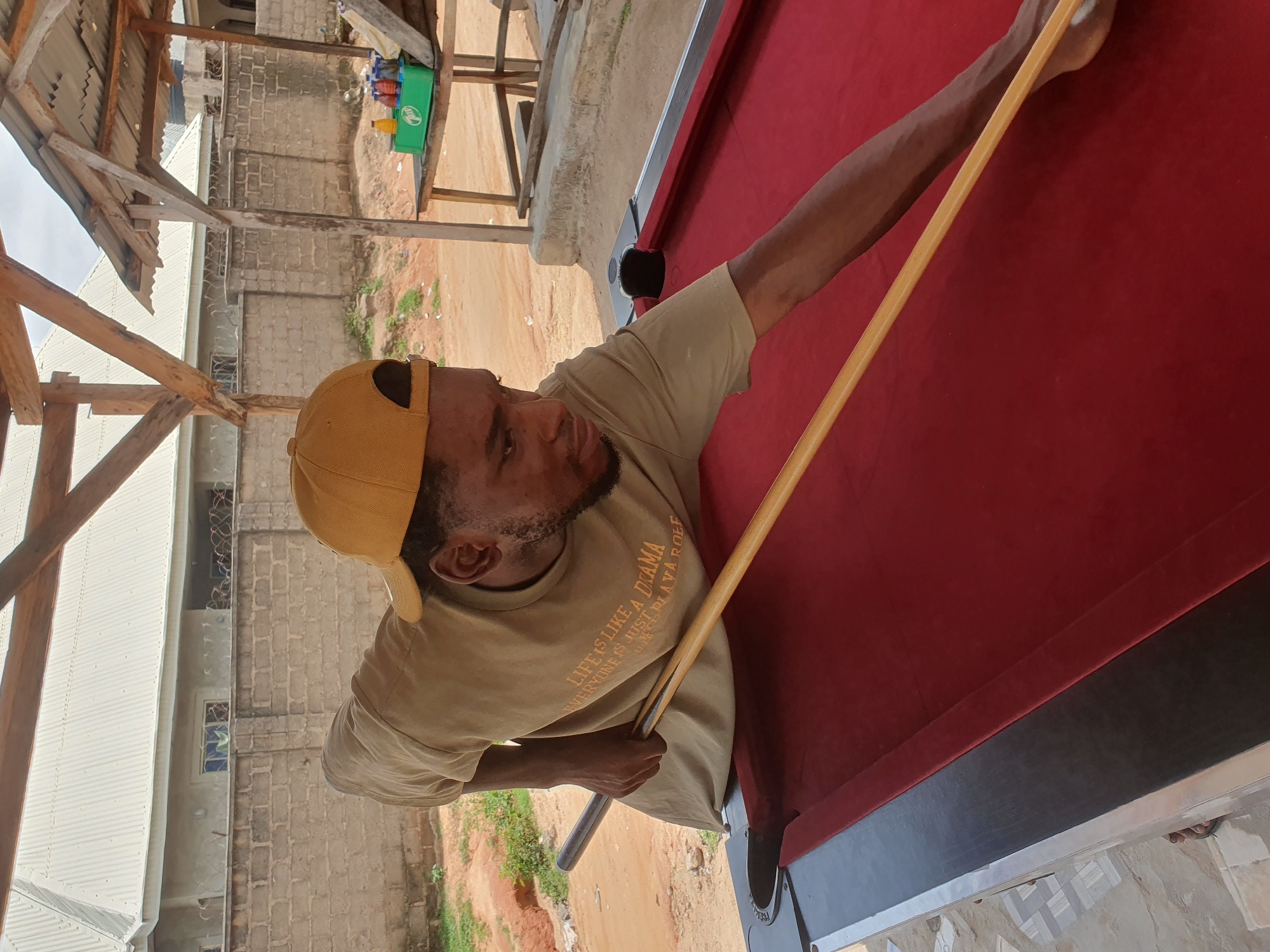 An adult male wearing a tan t-shirt with text and a yellow baseball cap plays pool. He is leaning over a red felt pool table, holding a pool cue with both hands, and appears to be taking a shot. The table is outdoors, underneath a simple wooden and corrugated metal shelter. In the background, there is a brick wall, a building with a white corrugated roof, and a sandy, unpaved area. The setting suggests a casual outdoor recreational space, possibly in Lokoja, Nigeria, given the architectural style and environment.