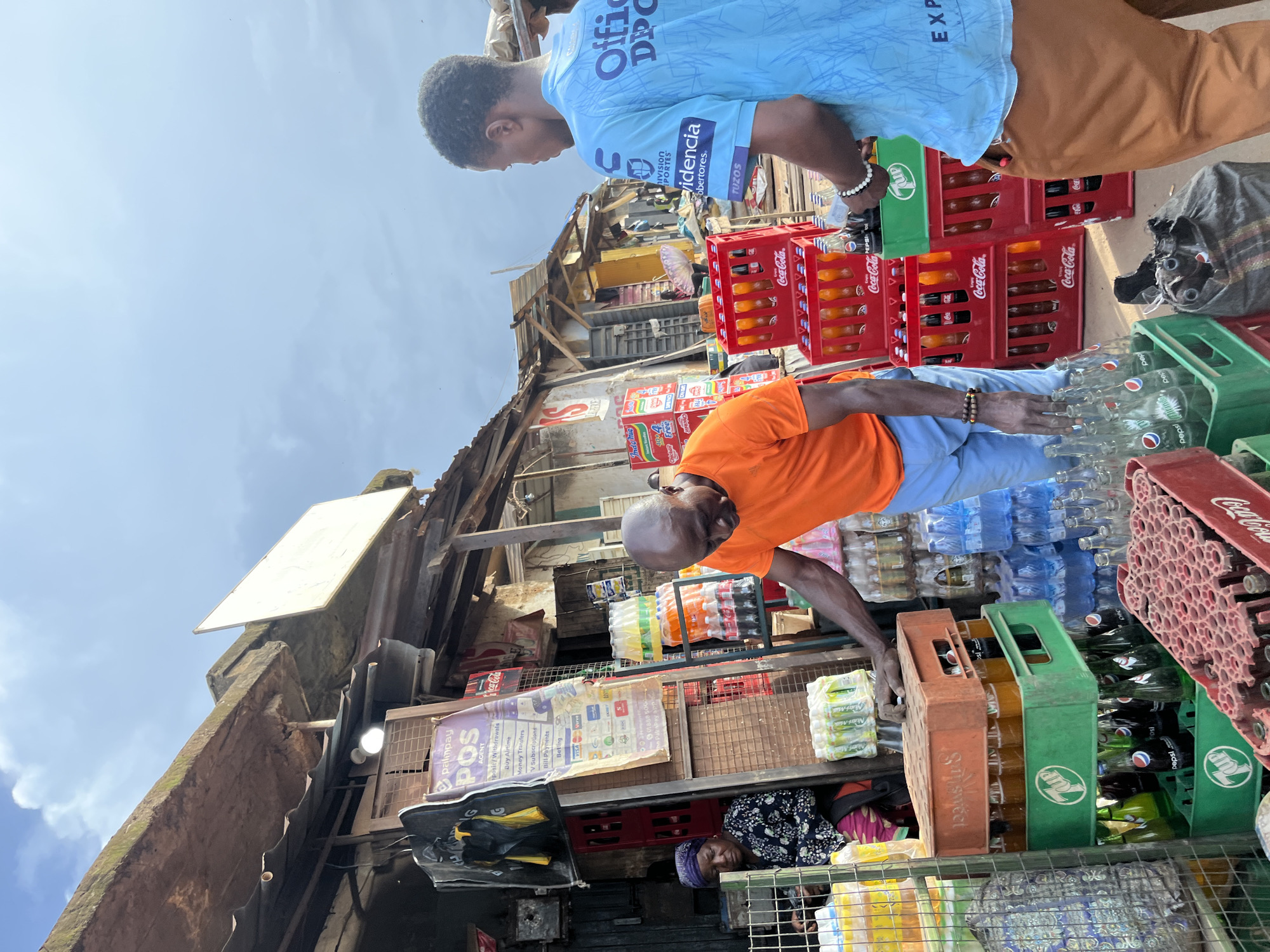 In the city of Rafin Par, Nigeria, an outdoor market scene unfolds under a cloudy sky. A man in an orange t-shirt and blue pants is seen reaching into a stack of crates filled with bottled beverages. Beside him, another man in a light blue t-shirt and brown pants also appears to be involved in handling the goods. In the background, a woman wearing a headscarf and a patterned top is seated, observing the activity. The market stall is cluttered with various crates of drinks, including Coca-Cola and Pepsi, as well as other packaged goods. A poster advertising "Palmpay POS Agent" is prominently displayed on the stall. The overall environment suggests a bustling commercial setting with a focus on the sale of drinks and related services.