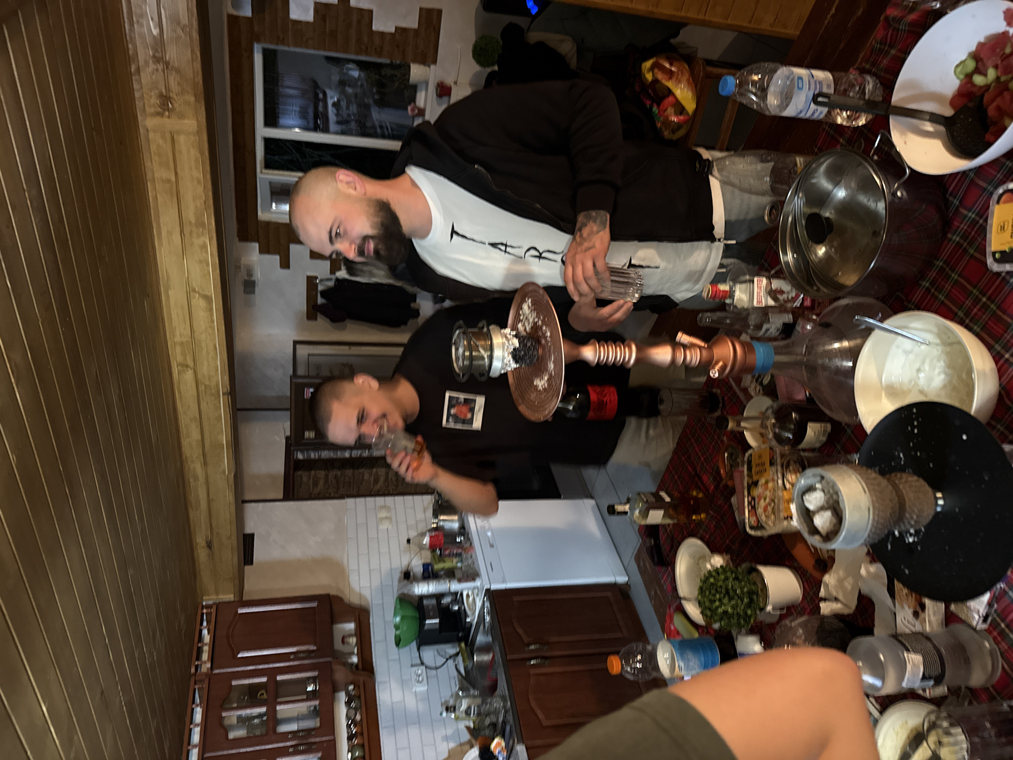 The scene is in what appears to be a kitchen and dining area. Two men are present; one is bald with a beard, wearing a white t-shirt and a black jacket, holding a glass. The other man has short hair and is wearing a black shirt. Both men are holding glasses.
A hookah is on the table, which is covered with a red plaid tablecloth. Other items present on the table include bottles of alcohol, water bottles, and bowls containing food.
Kitchen cabinets are visible in the background, along with a refrigerator. The location is suspected to be in Ribaritsa, Bulgaria.