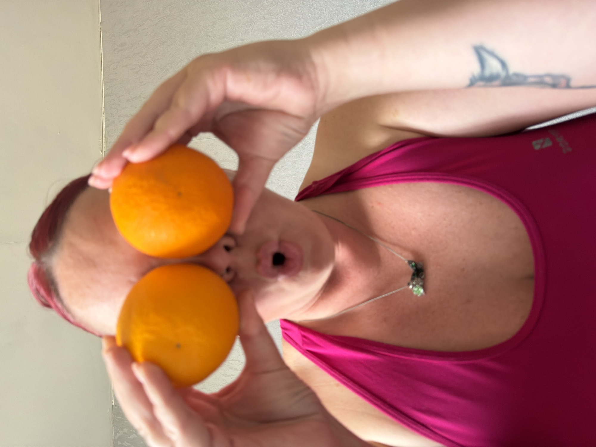 A rotated close-up shot shows a woman in a pink tank top holding two round, orange fruits in front of her eyes. Her hair is reddish-purple, and she wears a silver necklace. She is making a puckered face. A tattoo is visible on her arm. The background is a textured, light-colored wall. The location is not discernible from the image.