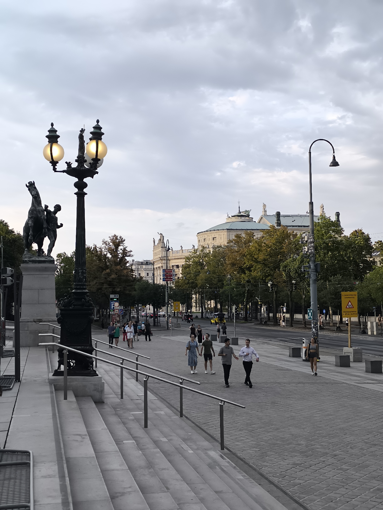 The image depicts a street scene in Vienna, Austria, under a cloudy sky. In the foreground, a wide set of stone stairs with metal railings leads up to a plaza. A large, ornate lamppost with two illuminated globes and a statue of a person riding a horse stands to the left of the stairs. The street itself is paved with cobblestones and is lined with trees. Several people are walking along the sidewalk. In the background, a grand building with a domed roof and classical architecture is visible, flanked by more trees and other buildings. Traffic is present on the street, including cars and a motorcycle. A yellow warning sign is positioned on the sidewalk to the right of the street. The overall atmosphere is that of a public thoroughfare in an urban European setting.