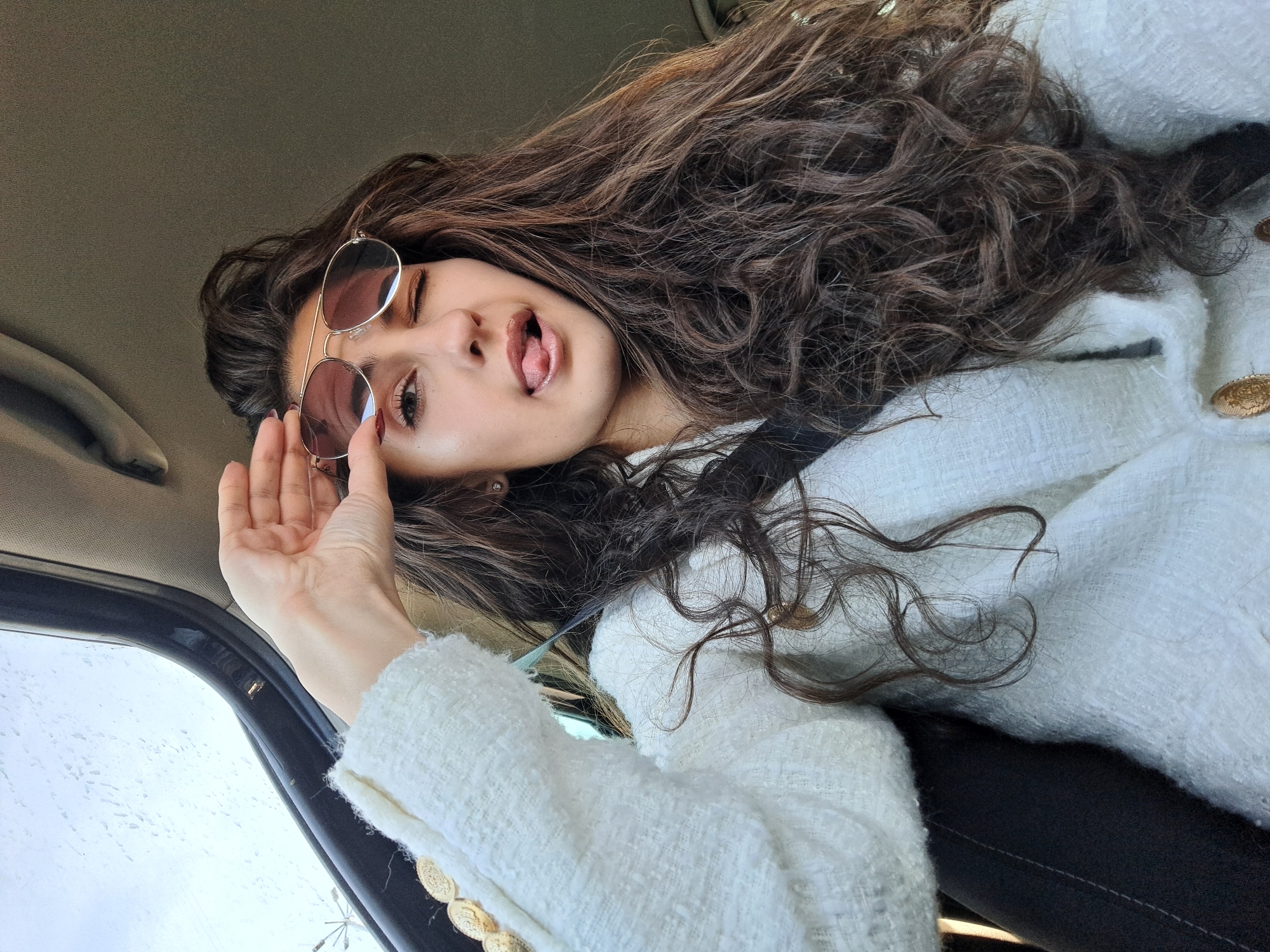 A woman with long, brown, curly hair is reclining in the passenger seat of a car. She is wearing a white tweed jacket with gold buttons and a seatbelt across her chest. She has round, gold-framed sunglasses on, which she is holding with her left hand, slightly obscuring her left eye as she winks. Her tongue is sticking out slightly. The car door and window are visible in the lower left corner of the image, and the sky can be seen through the window. The location is Unknown, Unknown.