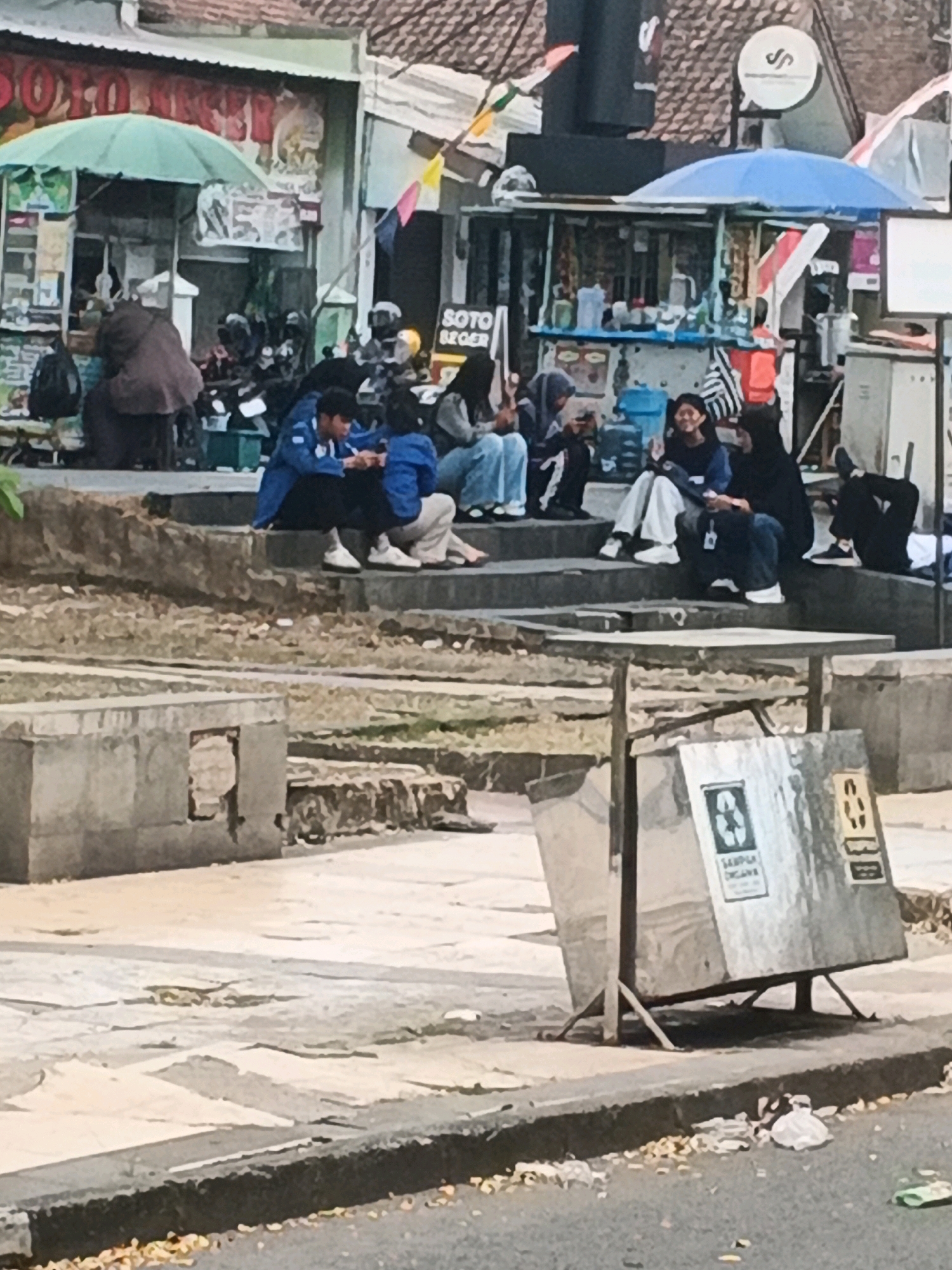 The image depicts an outdoor scene in Kalirejo, Indonesia, likely a public gathering space or marketplace. In the foreground, a silver metal trash receptacle with recycling symbols and Indonesian text is positioned at the edge of a paved area. The receptacle is tilted and supported by a metal frame, with scattered litter around its base. Behind the receptacle, a tiered concrete structure serves as seating for a group of young people.
Several individuals are seated on the concrete steps, engaged in conversation or observing their surroundings. Some are wearing casual clothing, including jackets and pants. To the left and background, food stalls or small businesses are visible, indicated by awnings, umbrellas, and displayed goods. A person in a brown garment is seen seated near one of the stalls. The overall environment suggests a bustling urban area.