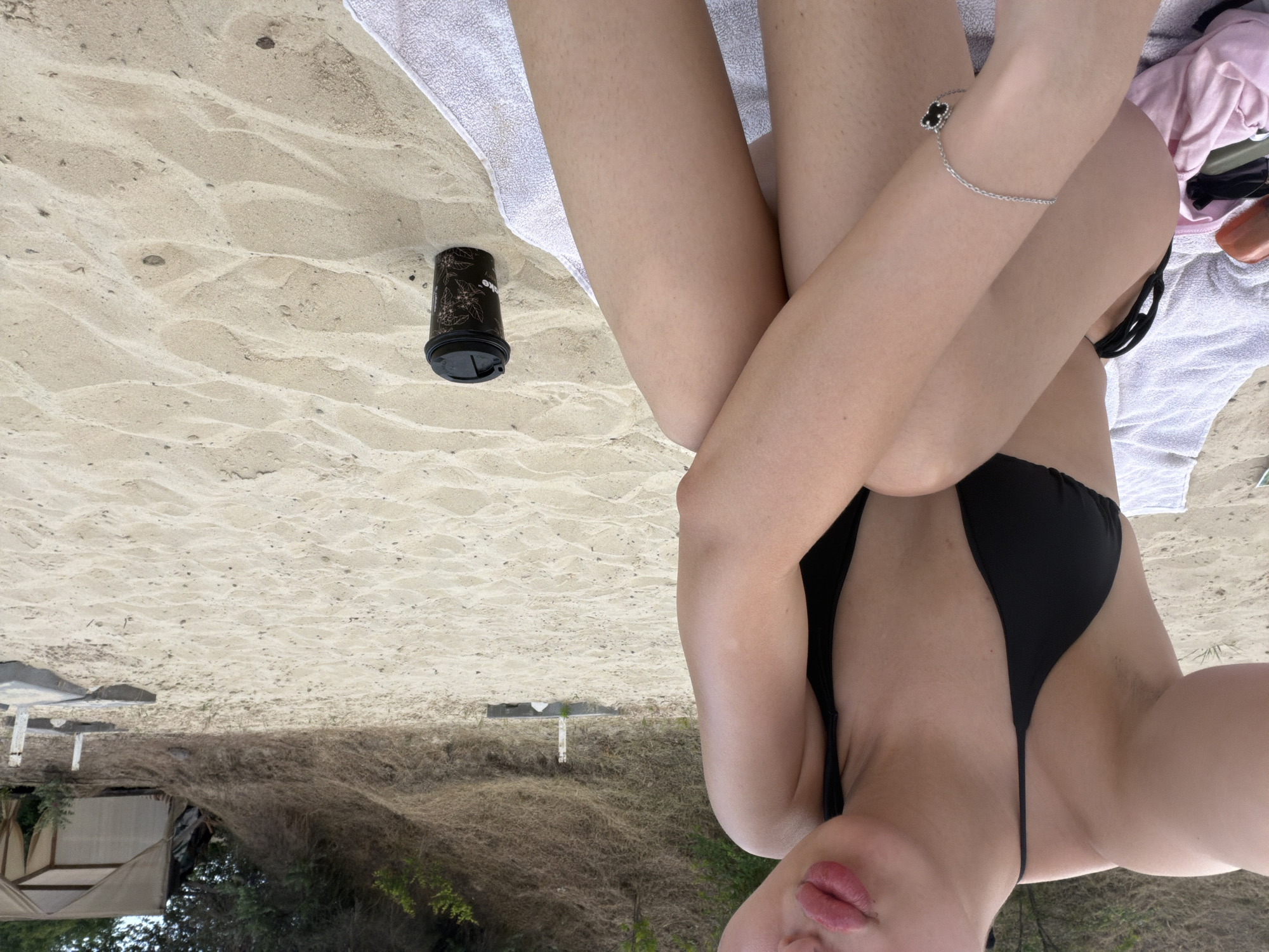 The image shows a person sitting on a beach. The person is wearing a black bikini and a silver bracelet with a black clover charm. They are sitting on a white towel, with their legs crossed. A black coffee cup with a floral pattern is placed on the sand next to them. In the background, there is a sandy hill with some dry vegetation and trees. Further in the distance, there is a tent-like structure. The scene appears to be a relaxed day at the beach.