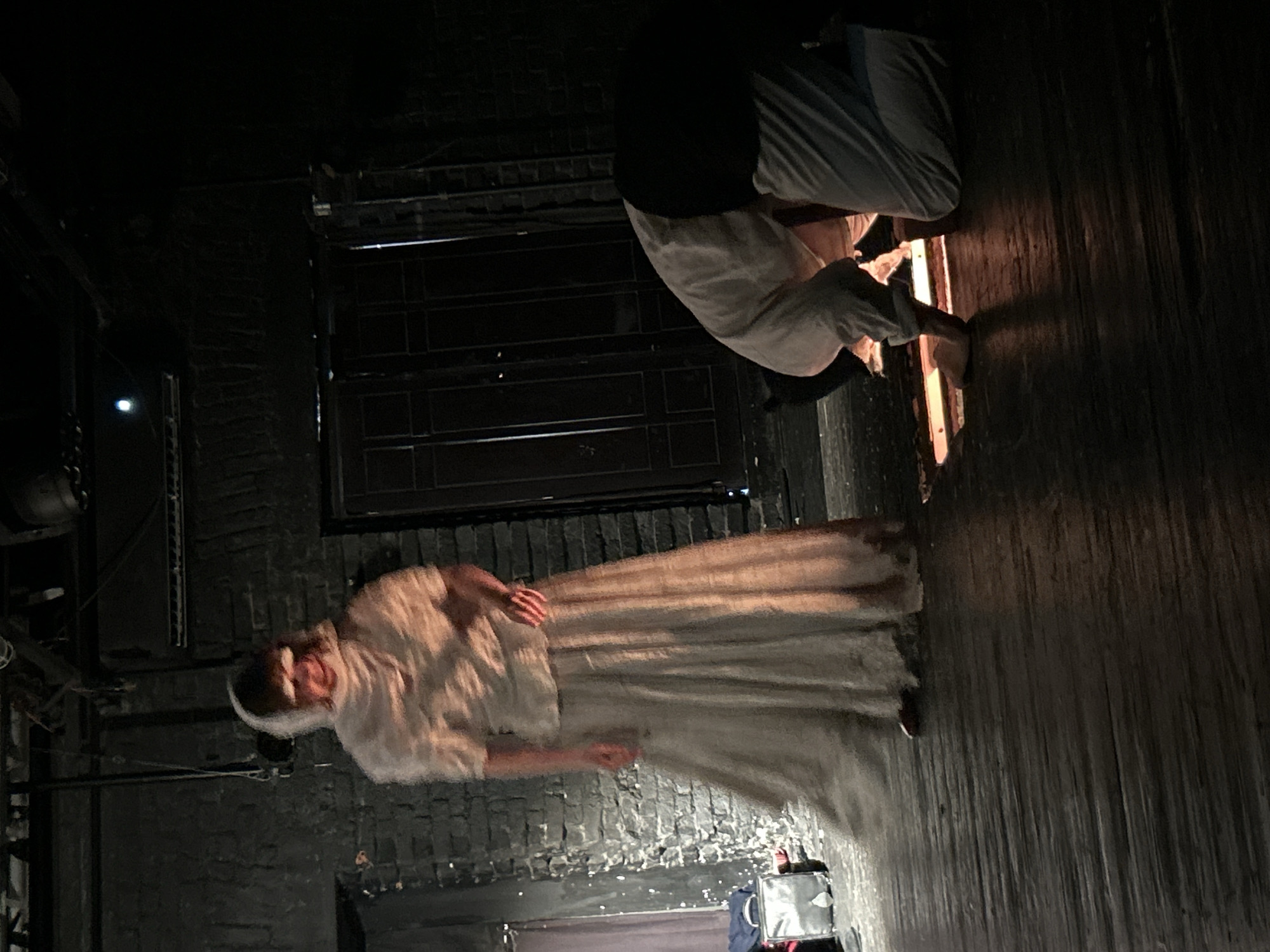 A dimly lit, unidentifiable indoor space features a person in a long, light-colored garment and head covering, possibly a theatrical costume. To the upper right, another individual is crouched down, wearing dark pants and a light-colored shirt. The floor appears to be wooden. A dark window or door is visible in the background. The scene is of indeterminate location and city.