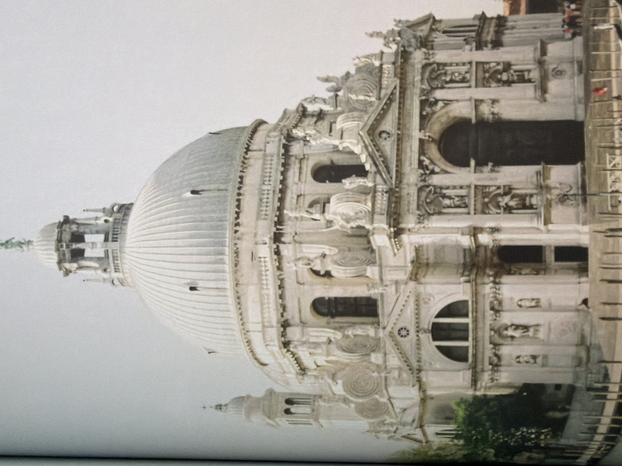 The image shows the Basilica di Santa Maria della Salute, a Roman Catholic church in the Dorsoduro district of Venice, Italy. The church is notable for its octagonal plan and its large dome, which is a prominent feature of the Venetian skyline. The building is made of white marble and is decorated with sculptures and other architectural details. The church is situated on the Grand Canal, and in the foreground, there is a body of water with a wooden fence and a few small boats. There are also a few people visible in the distance. The sky is overcast.