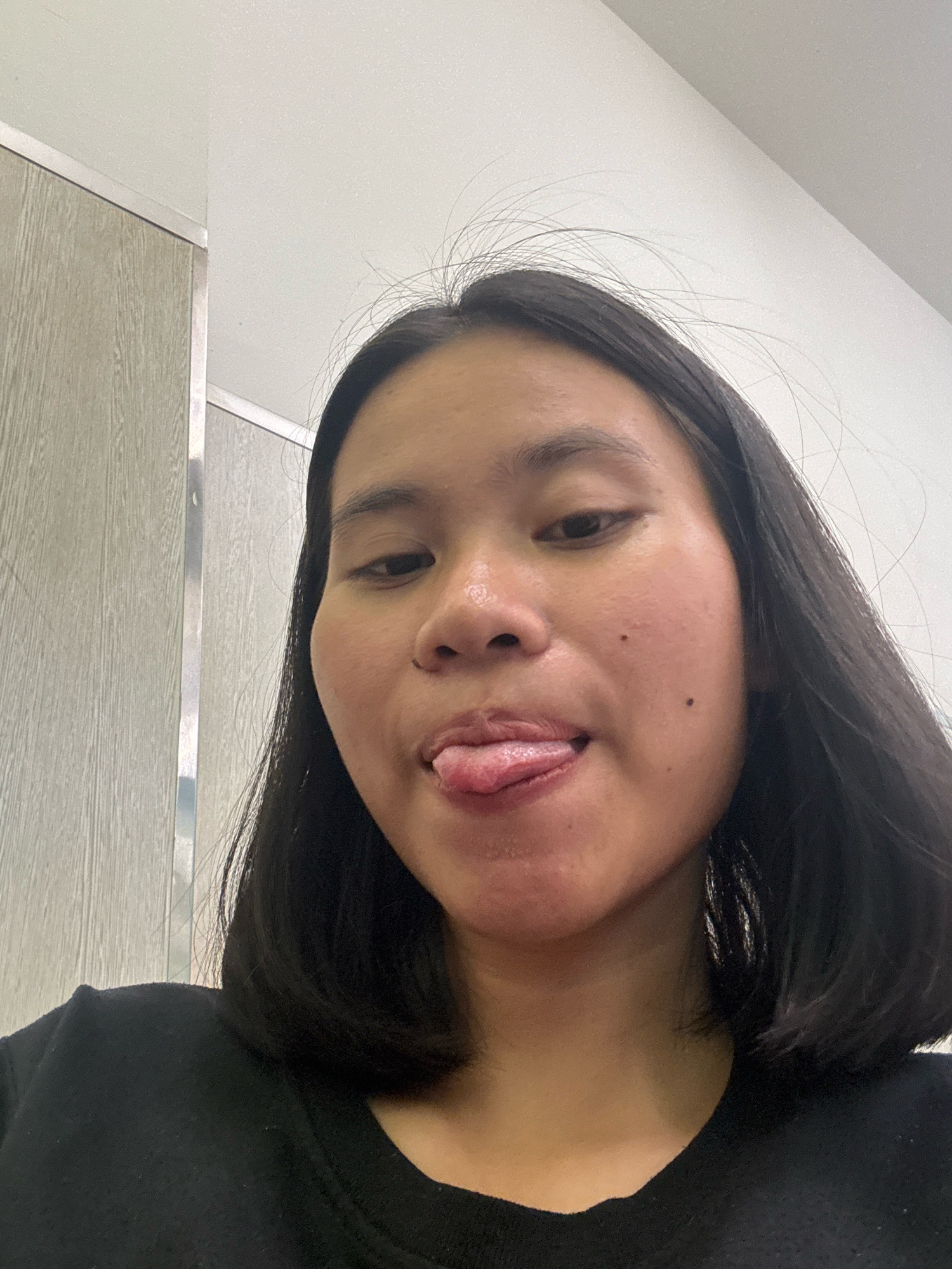 A young woman with dark hair is pictured indoors, sticking her tongue out. She is wearing a black t-shirt. The background consists of a light-colored wall and a wooden paneling that appears to be part of a cabinet or door. The lighting is bright, suggesting an indoor setting. The image was taken in East Jakarta, Indonesia.