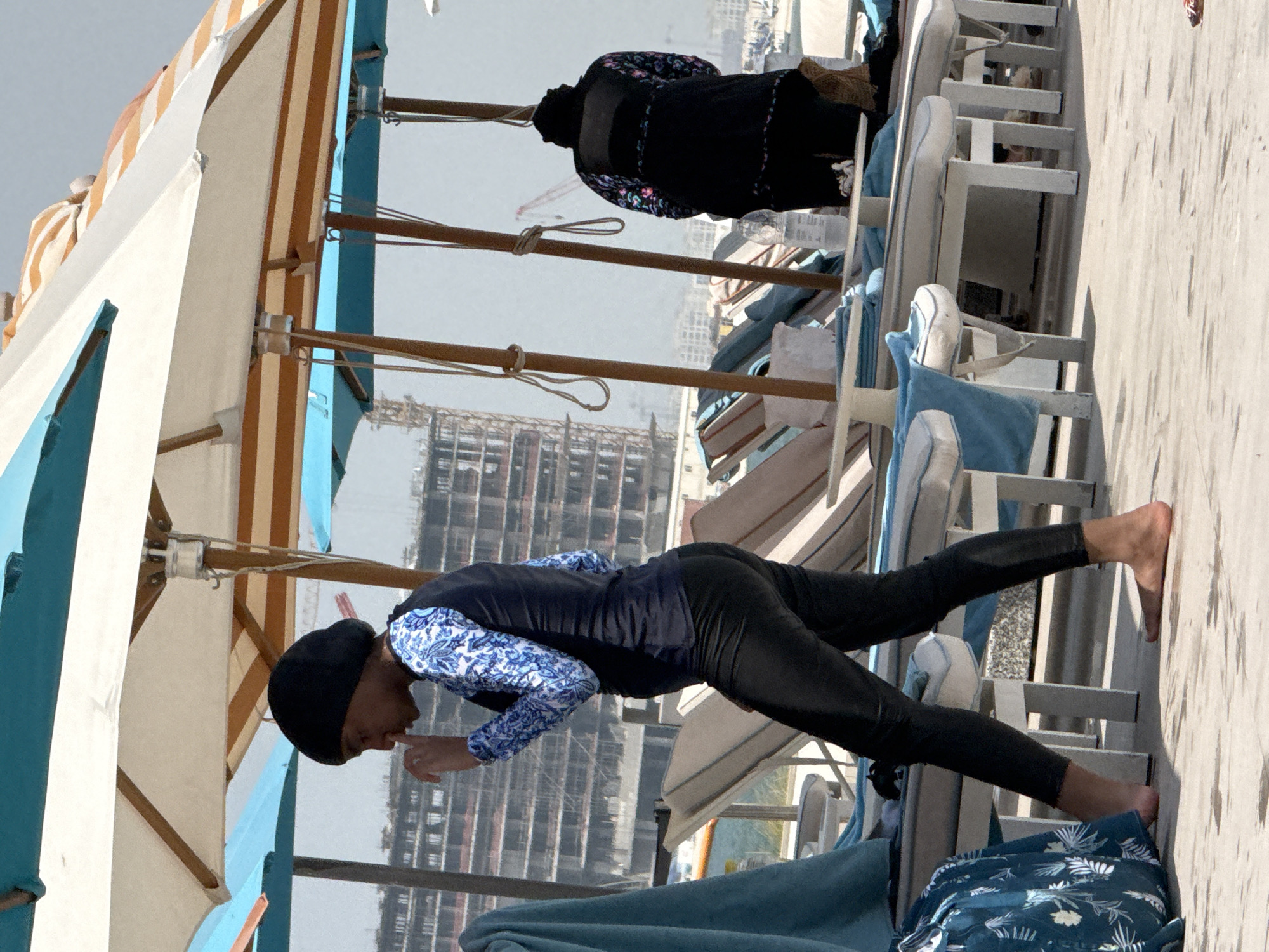 The image depicts a sunny day at a beach resort in Ras Al-Khaimah, United Arab Emirates. In the foreground, a person wearing a black headscarf and a long-sleeved rash guard with blue and white floral patterns is walking. They are also wearing black leggings. The person is barefoot and their right index finger is near their mouth. Behind them, another person is standing, wearing a black headscarf and a black garment with colorful floral patterns on the sleeves and trim. They are also barefoot.
The background shows beach lounge chairs with blue towels draped over them, and several beach umbrellas providing shade. Beyond the beach area, construction is underway on a large building, with cranes visible in the distance. The overall environment suggests a busy resort with ongoing development.
