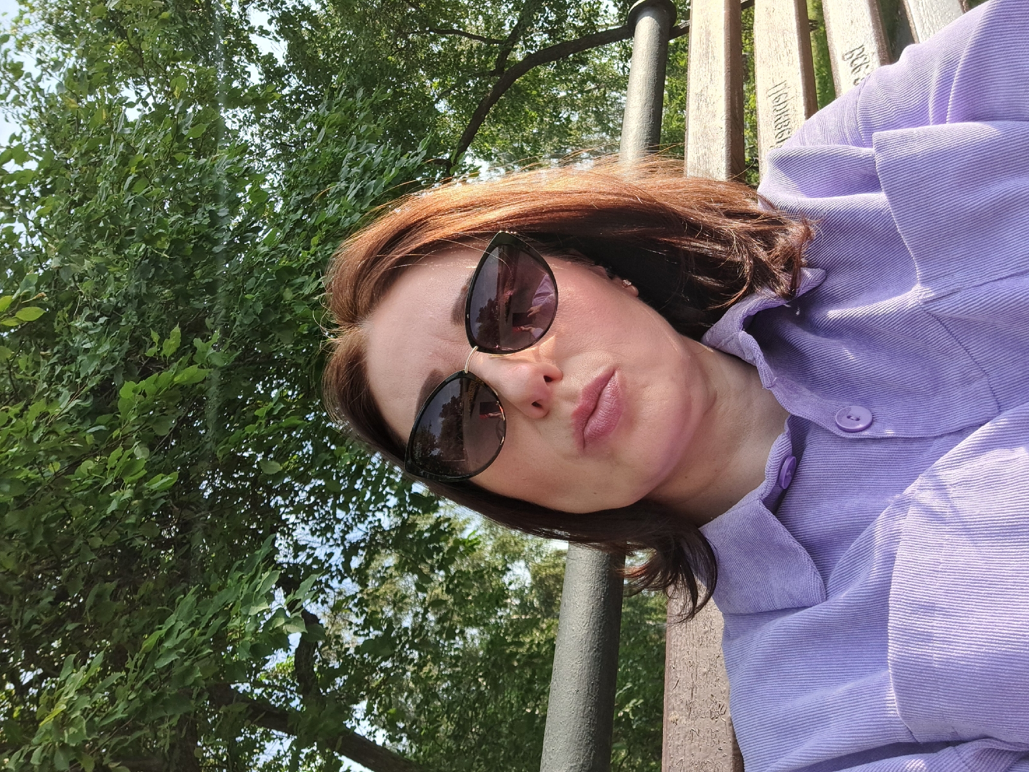 The image shows a woman with auburn hair, wearing sunglasses and a lavender corduroy shirt. She is sitting on a wooden bench in what appears to be a park or outdoor public space, with green trees and foliage in the background. The lighting suggests it is daytime. There is no specific information in the image to indicate the location is the city of Irkutsk, Russia.