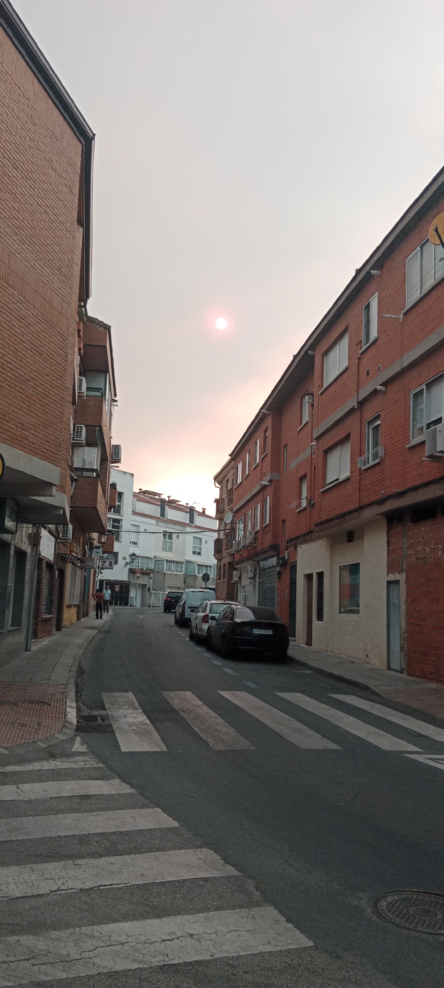 This is a vertical, eye-level shot of a residential street in Galapagar, Spain. The sun, a faint pinkish orb, is visible in the hazy sky, casting a soft light. The street is lined with multi-story brick buildings on both sides. On the left, the buildings are mostly brick, with some white and light-colored sections. Air conditioning units are visible on balconies. On the right, the buildings are primarily red brick with white window frames and shutters. The street has a pedestrian crossing marked by white stripes. Several cars are parked along the right side of the street, including a black sedan with a license plate reading "B145 CCD." Two individuals are standing on the sidewalk on the left side of the street near the entrance to a building. The overall atmosphere is quiet and somewhat overcast.