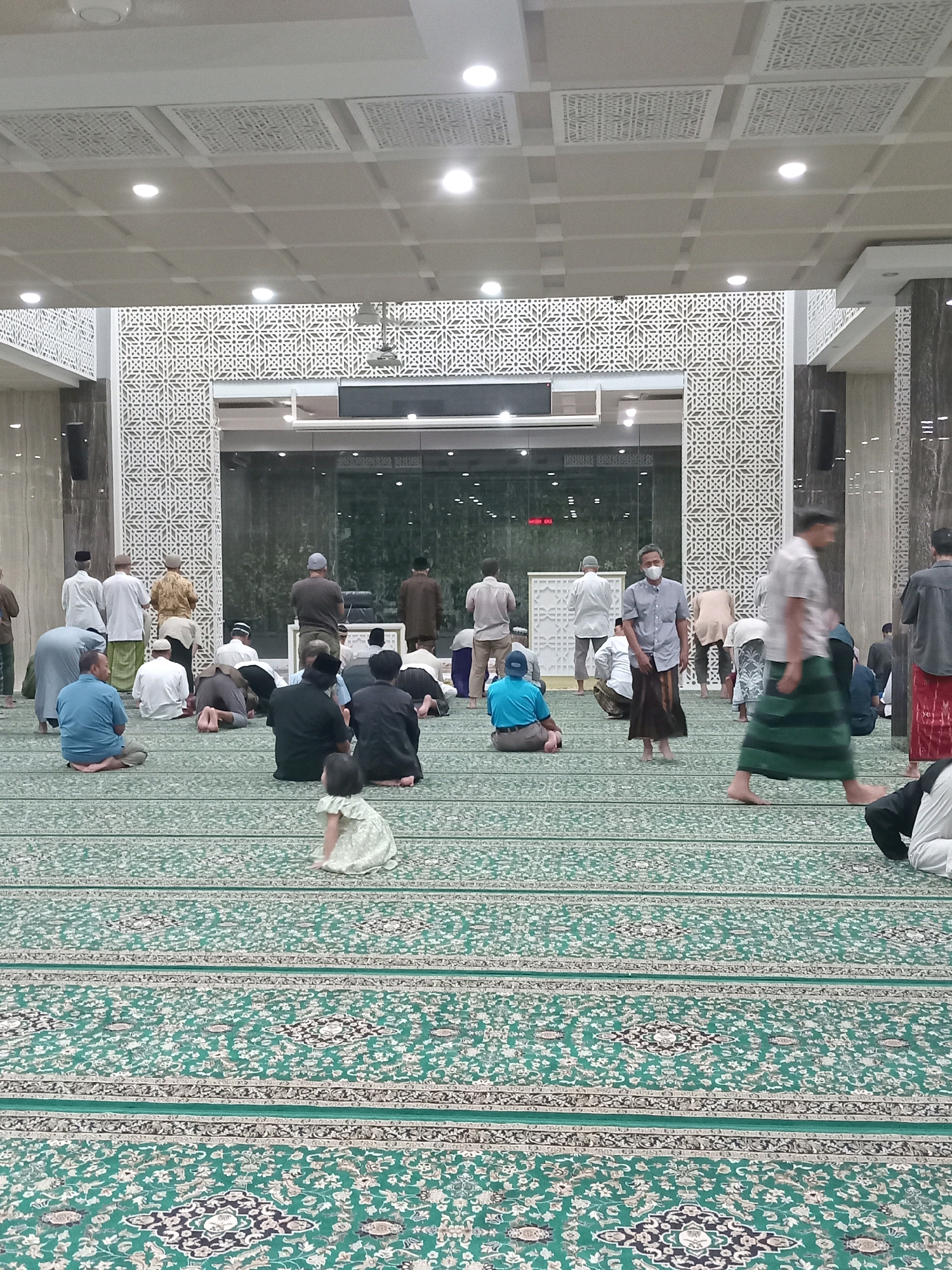The image depicts the interior of a mosque in Randuagung, Indonesia, likely during prayer. A large number of people are present, most of whom are men, dressed in traditional Islamic attire, including long shirts, sarongs, and caps. Several individuals are kneeling or prostrating on a green carpet adorned with intricate patterns, indicating they are engaged in prayer. Some people are standing, possibly waiting to join the prayer or observing. A young child in a light-colored dress is visible in the foreground, sitting on the carpet. The mosque's architecture features decorative geometric patterns on the walls and ceiling, with modern lighting fixtures. The overall atmosphere appears serene and communal, with people participating in a religious observance.