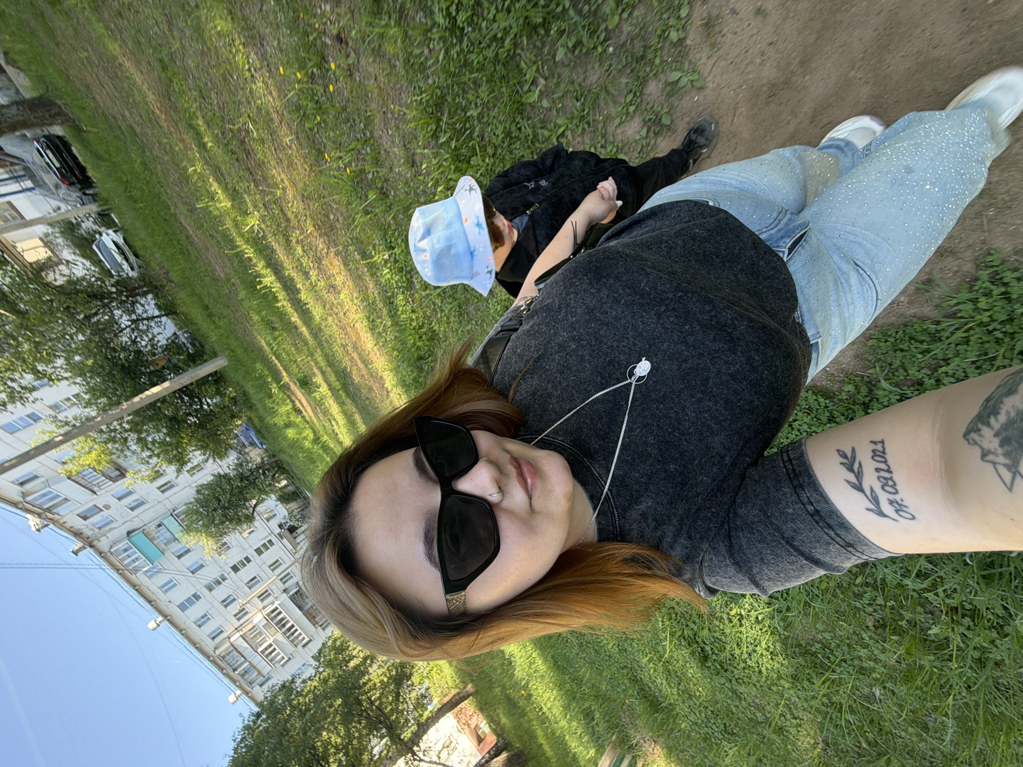 A selfie depicts a woman outdoors with a child. The woman, positioned closest to the camera, wears dark sunglasses, a dark t-shirt, and bedazzled light blue jeans. She has light brown hair with darker roots and a tattoo on her right forearm. A child in a black jacket and blue bucket hat is next to her. They are on a grassy area with a path next to it. In the background, there are apartment buildings. The image was taken in Chaikovsky, Russia.