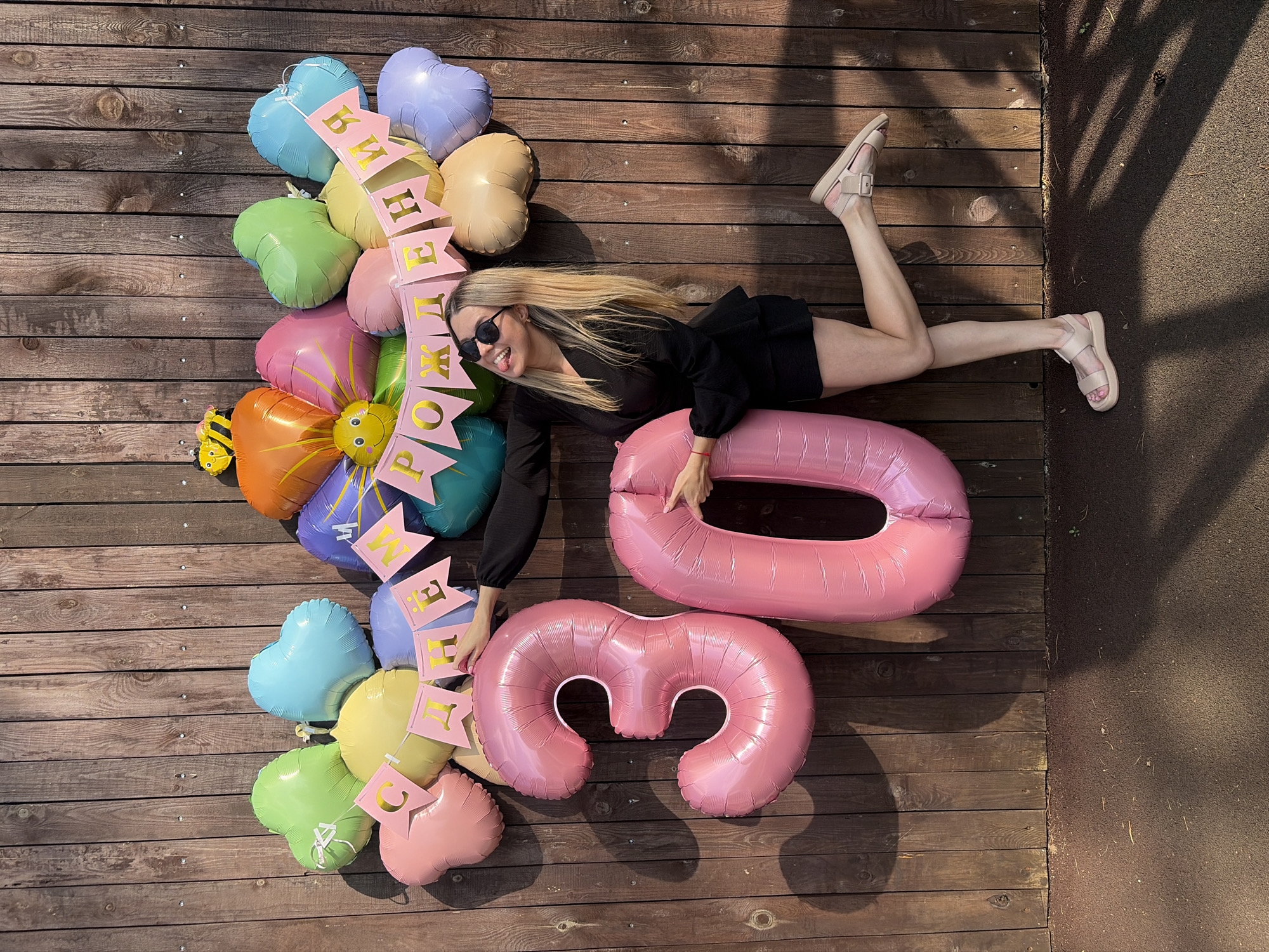 A blonde woman in a black outfit and sunglasses is posing with large pink balloons shaped like the numbers 3 and 0. She is standing in front of a wooden fence, with a garland of heart-shaped balloons and a banner that reads "С ДНЕМ РОЖДЕНИЯ" (Happy Birthday) in Russian. The scene appears to be outdoors, possibly in a yard or park, given the wooden fence and a shadow pattern on the ground. The woman is playfully sticking out her tongue and looking towards the camera, with her right leg bent at the knee and extended backward. The balloons are arranged in a celebratory manner. The overall impression is a cheerful birthday celebration. There is no indication of the city of Chaikovsky, Russia, in the image itself.