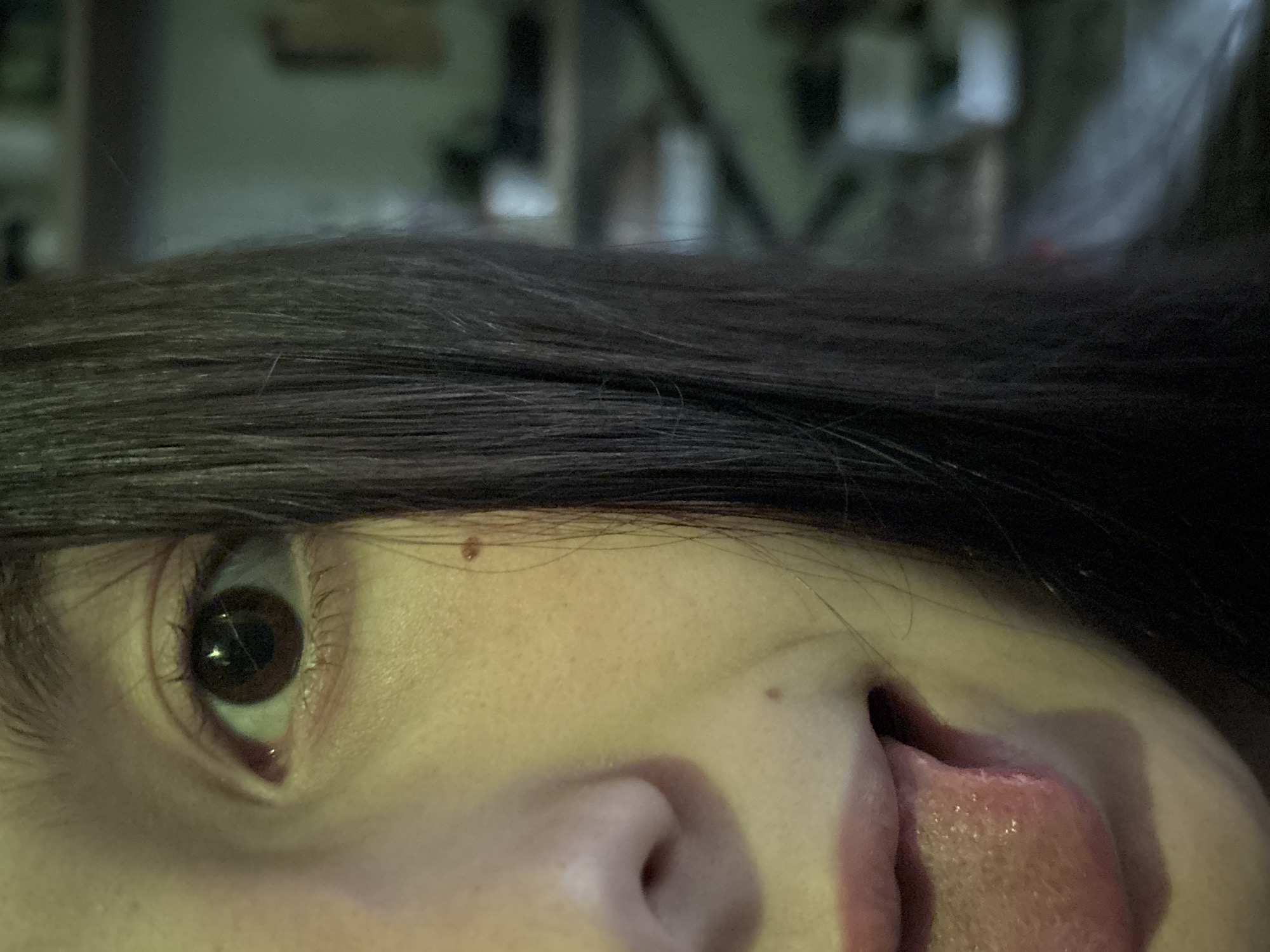 This is a close-up, waist-up, eye-level shot of a person's face, focused on the left side of their face and their tongue. The background is out of focus, showing shelves with various objects. The image is brightly lit with a yellow tint. The person has brown eyes, dark hair, and two moles on their face. Their mouth is open, and their tongue is sticking out, showing a moist, pink surface. The image does not provide any information about the location in the city of Фоки, Russia.
