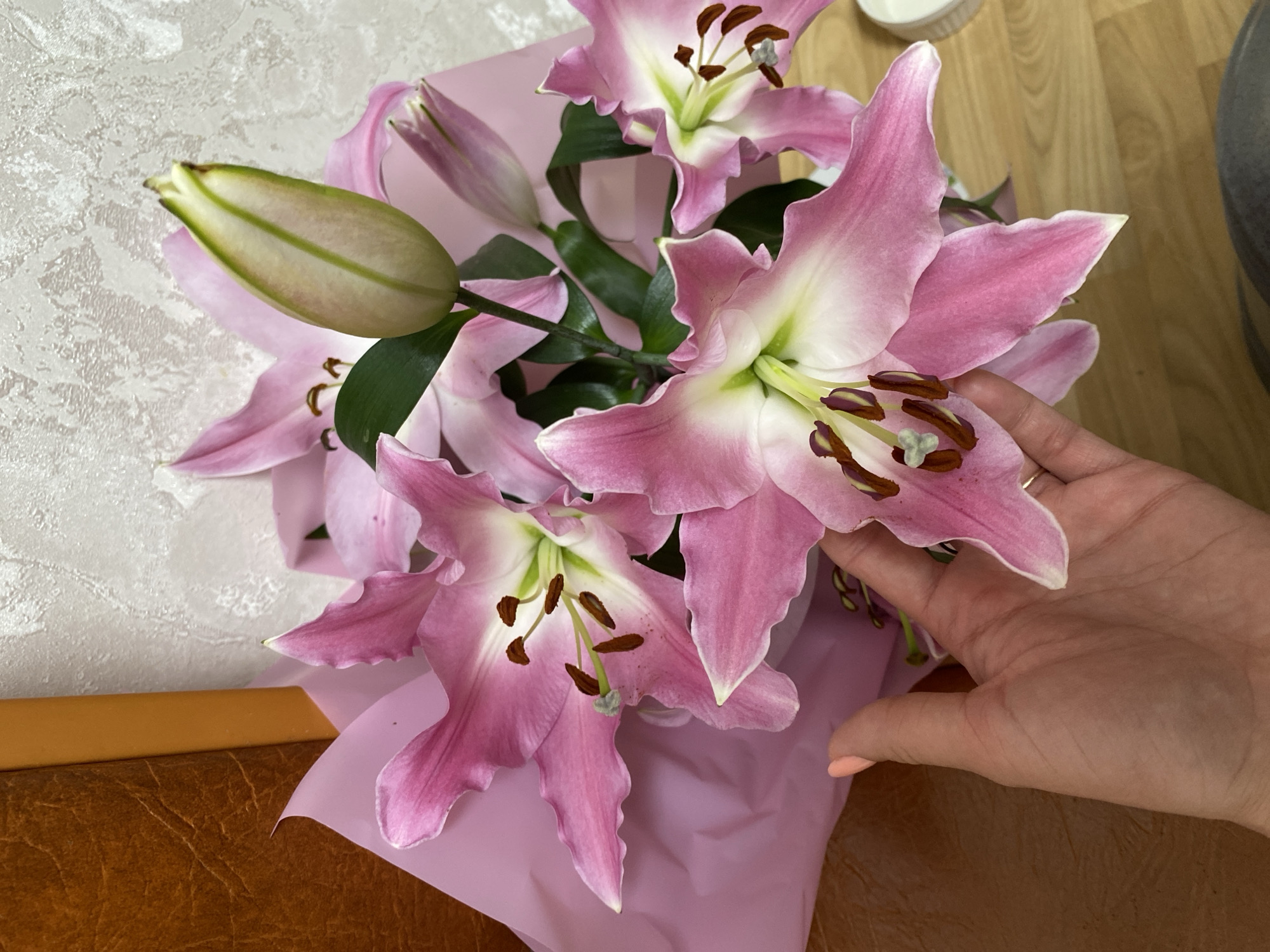 A bouquet of pink and white lilies wrapped in light pink paper is arranged on what appears to be a brown surface, possibly a table or counter. A person's hand with a pale pink nail polish is visible near the flowers, possibly adjusting or holding them. The background includes a light-colored patterned wall and a glimpse of wooden flooring. The location is presumed to be Chaikovsky, Russia.