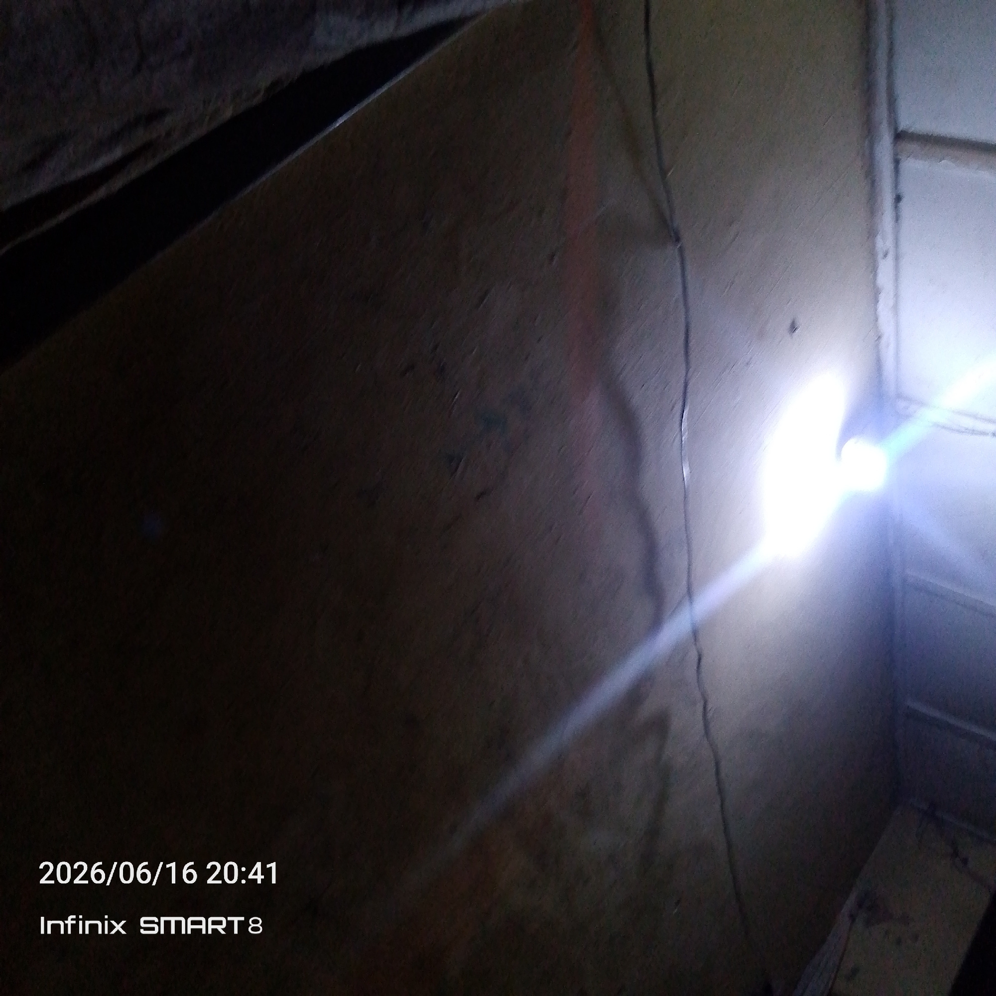 The image captures an interior scene, likely a dimly lit room or storage area. A significant portion of the frame is dominated by a textured, light-colored wall with visible imperfections and a faint reddish line running vertically. A bright light source, possibly a flashlight or camera flash, illuminates the wall and casts a strong glare and lens flare across the right side of the image. In the lower left corner, a timestamp "2026/06/16 20:41" and device information "Infinix SMART 8" are visible, indicating the date and time the photograph was taken. The scene appears to be located in Jos, Nigeria, although no specific landmarks or contextual clues within the image itself confirm this location. The overall impression is of a utilitarian or neglected space, with the light creating a stark contrast against the shadowed areas.