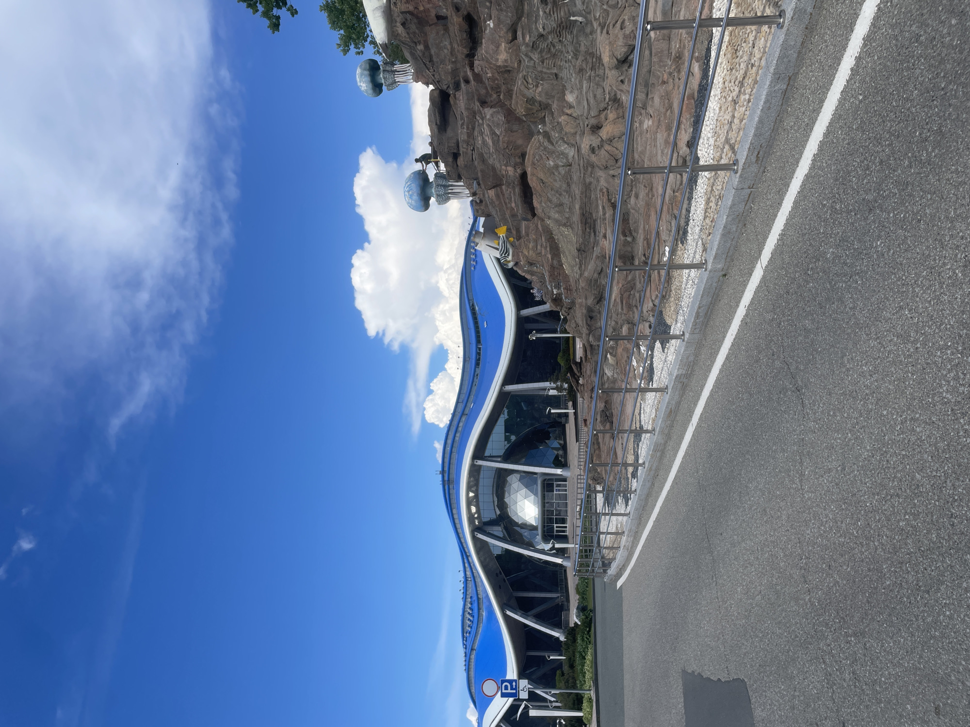 The image depicts the Primorye Oceanarium on Russky Island, Vladivostok, Russia, under a bright blue sky with scattered white clouds. The foreground shows a paved road curving to the left, bordered by a white line and a low, grey concrete curb. A metallic railing runs along the curb, separating it from a rock formation. The rock formation is composed of large, rough-hewn brown rocks, with a decorative element resembling a blue jellyfish on a white pedestal at the top. A person is visible on the rock formation, interacting with the jellyfish sculpture. Further to the left, the oceanarium building is prominently featured. It has a distinctive undulating blue roof with white structural supports. The facade is largely glass, revealing the interior structure. A blue and white sign with a "P" for parking and an accessibility symbol is visible near the road. The overall scene suggests a public space with architectural and artistic elements.