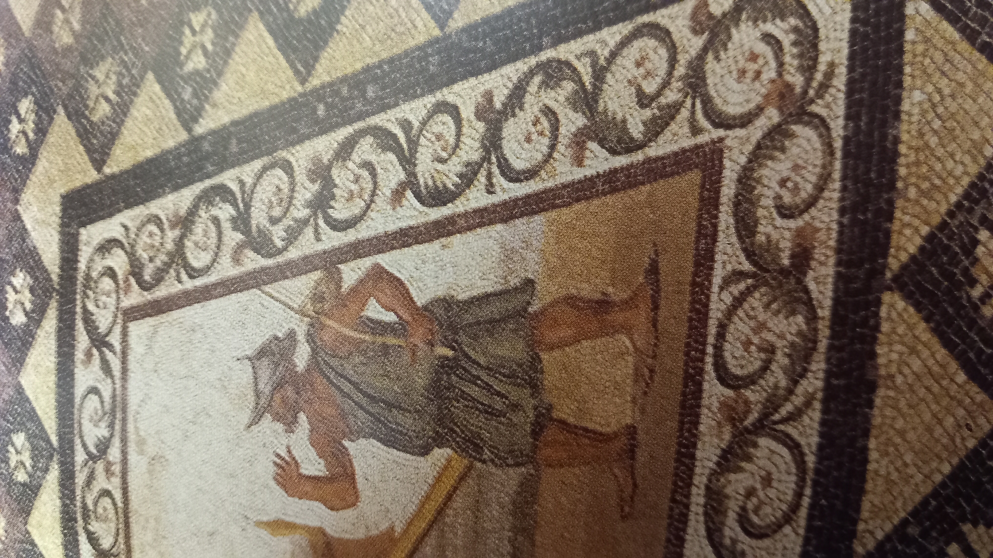 The image displays a mosaic floor depicting a man in ancient attire. He wears a helmet and a draped garment, and is holding a bow and arrow. The mosaic is part of the Casa Romana, a Roman-era villa located in the city of Kos, Greece. The floor features intricate geometric patterns and a decorative border surrounding the central figure. The colors used are earthy tones of brown, beige, and white, characteristic of ancient mosaics.