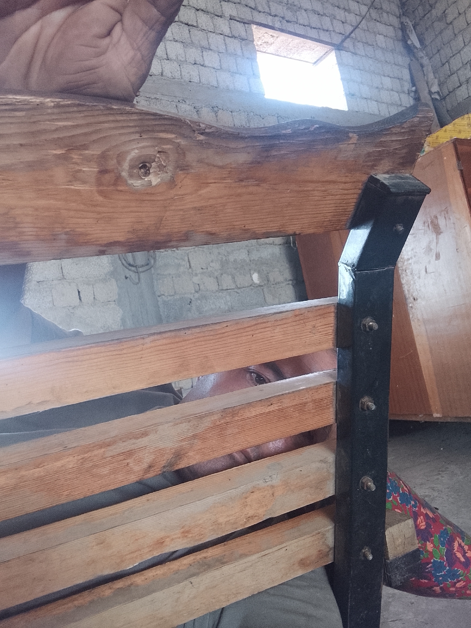 The image captures a close-up view of a partially constructed wooden structure, likely a bed frame, in an interior setting. The dominant elements are aged, weathered wooden planks and a black metal support frame. Light emanates from a small, open window high on a brick wall, illuminating the scene. A person's eye is visible through a gap in the wooden slats, suggesting an interaction or presence within the structure. The background shows unfinished brick walls and what appears to be a piece of patterned fabric. The scene suggests a location undergoing renovation or being used as a workshop. The specific location in Sabir Rakhimov, Uzbekistan, cannot be determined from the visual information alone, but the style of construction and materials are consistent with the region.