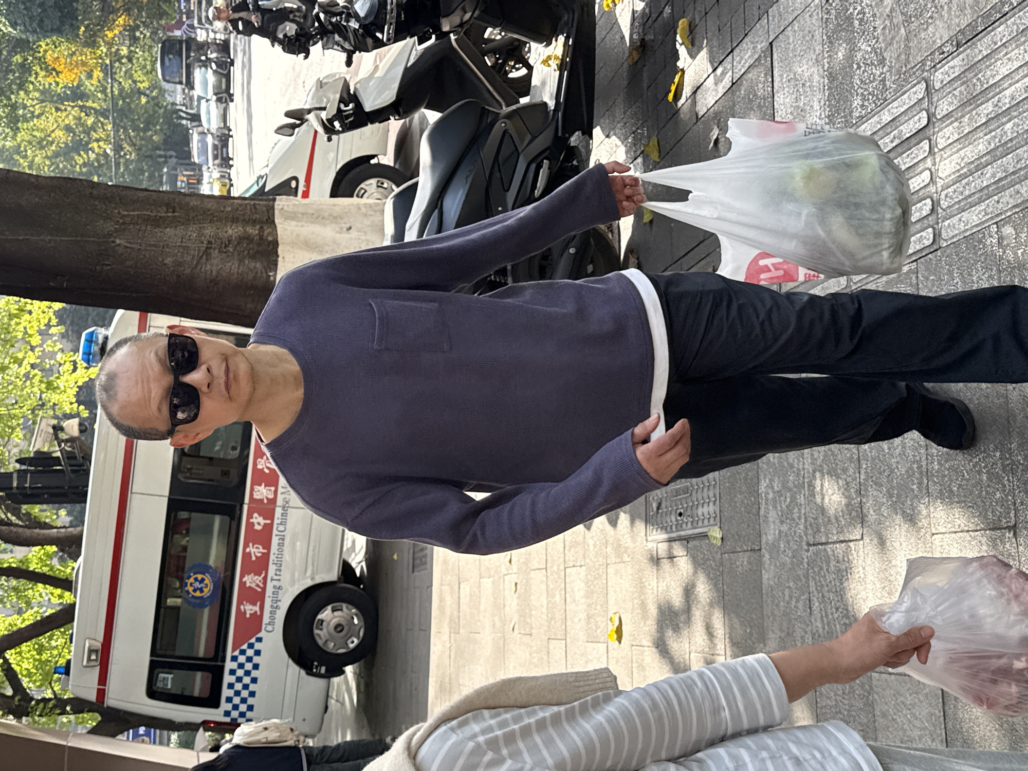 A street scene in Yuzhong District, China, shows a man standing on a sunlit sidewalk, holding a white plastic bag. The man is middle-aged, wearing black sunglasses, a dark purple long-sleeved shirt over a white undershirt, and dark trousers with black shoes. His facial expression is not discernible due to the sunglasses. His right hand holds the white plastic bag, which appears to contain some light-colored items. In the immediate foreground, another person's hand is visible, holding a similar transparent plastic bag that contains what looks like red meat. This suggests an exchange of goods or a recent purchase.
The background features a bustling urban environment. A large white service vehicle, resembling an ambulance or medical transport van, is parked behind the man. It has blue lights on the roof, a red and blue striped design, and a blue and white checkered pattern along its lower side. Visible text on the side of the vehicle reads "重庆市中医医" (Chóngqìng Shì Zhōngyī Yī - Chongqing Traditional Chinese Medicine) and below it, "Chongqing Traditional Chinese M". Further down the street, multiple motorcycles are parked, and other vehicles can be seen in the distance. Lush green trees line the street, with some autumn leaves scattered on the ground. The weather is clear and sunny, casting distinct shadows on the paved sidewalk, indicating daytime.