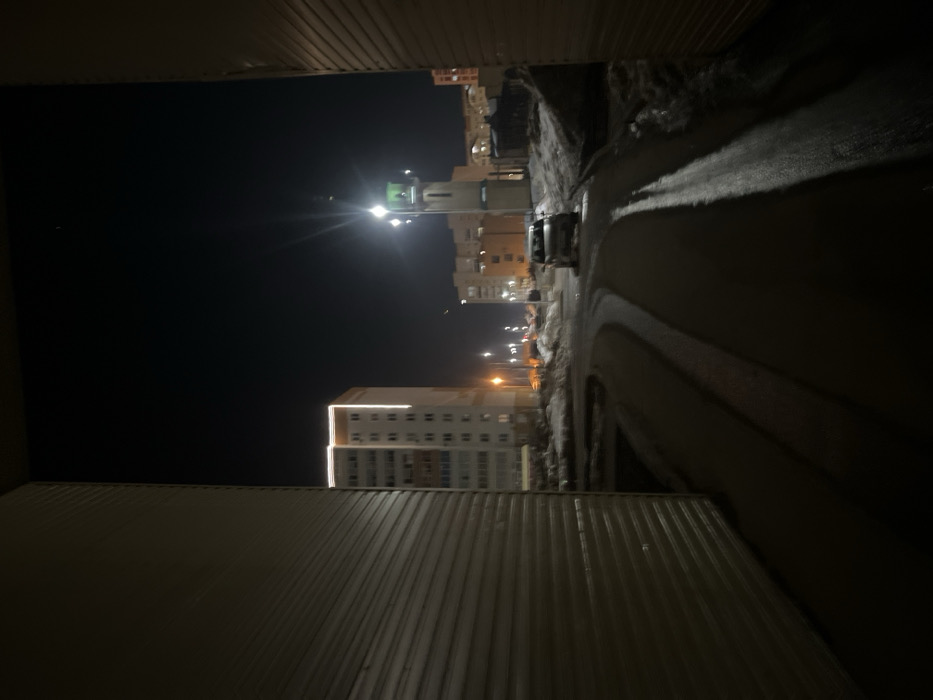 The image depicts a nighttime scene in a residential area of Yelabuga, Russia, captured from a narrow opening between two large, light-colored corrugated structures. A dark, wet or icy asphalt road curves from the immediate foreground into the middle distance, reflecting artificial lights. Patches of snow and dirty ice are present along the road's edges and on elevated terrain.
In the mid-ground, a light-colored sedan or hatchback is visible on the right side of the road, oriented away from the viewer. Further down the road, multiple streetlights with orange and white illumination sources are active. A prominent, tall, slender minaret with a light-colored shaft and a distinct green, conical cap stands to the right of the road. Bright light emanates from the minaret's upper section, creating a starburst effect.
In the background, several multi-story residential buildings are visible. A tall, light-colored apartment building on the left features illuminated white light strips along its top edge and vertical corners. Other residential structures with illuminated windows are visible further in the distance. The sky is dark. No individuals are visible, and no discernible actions or interactions are occurring. The minaret's architecture suggests a modern mosque complex, likely the Jami Mosque or a similar contemporary religious structure within a Yelabuga residential district.