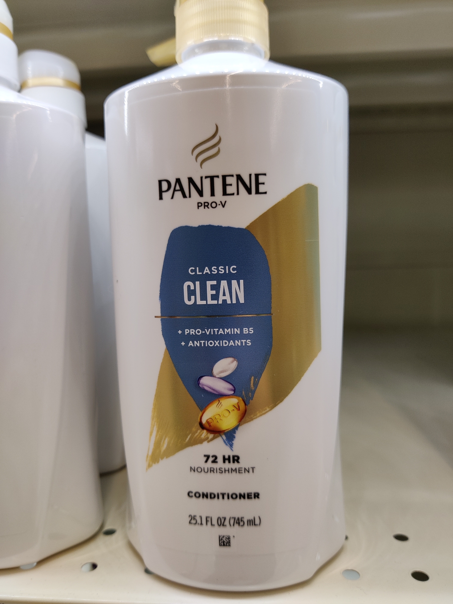 A close-up, eye-level shot shows the front of a Pantene Pro-V Classic Clean conditioner bottle. The bottle is white with a gold pump dispenser and a gold and blue label. The label features the Pantene logo, "PRO-V," "CLASSIC CLEAN," "+ PRO-VITAMIN B5 + ANTIOXIDANTS," and "72 HR NOURISHMENT." Below this, it reads "CONDITIONER" and "25.1 FL OZ (745 mL)." A QR code is visible at the bottom of the bottle. The bottle is placed on a shelf, and another white bottle is partially visible to its left. The background is a muted, neutral color. The image was taken in Mechanicsburg, United States of America.