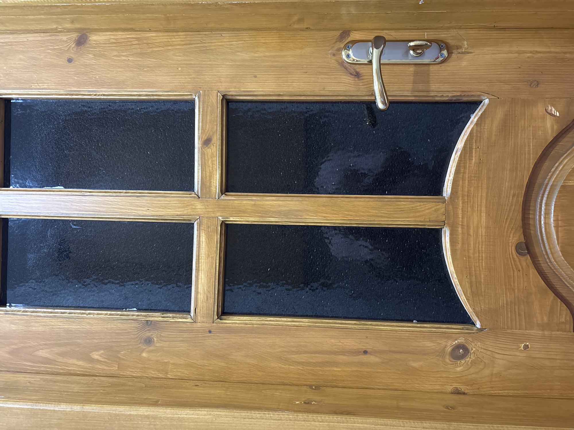 The image shows a wooden door with four rectangular panes of dark, textured glass. A gold-colored door handle is visible on the right side of the door. The door is constructed from light-colored wood planks with visible wood grain and knots. The lower portion of the door shows carved decorative elements. The scene is indoors or in a sheltered area, as indicated by the lighting and the clean appearance of the door. There are no people or animals visible in the image. The location of Novocherkassk, Russia is not discernable from the visual information provided in the image.