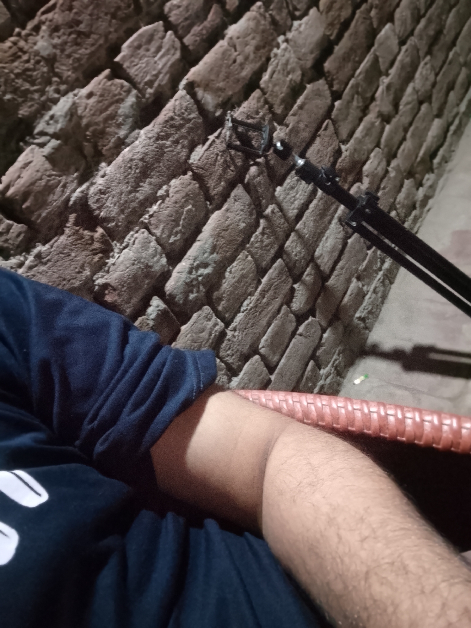 A person is seated, wearing a dark blue shirt with white markings. Their right arm is resting on an orange woven surface. Behind them is a textured brick wall. A black object, possibly a stand or tripod, is positioned near the wall. The location is Unknown, Unknown.