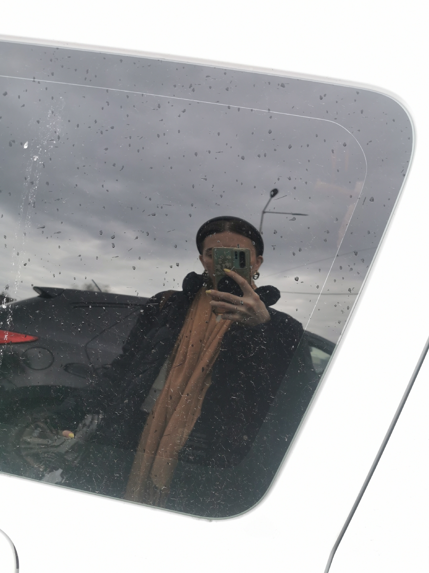 A woman, reflected in a rain-speckled car mirror, holds a phone with a light green case. She wears a black jacket and a light brown scarf or top, and a dark cap or headband. Outside the car, a dark gray SUV is visible, along with a street lamp and an overcast sky. The vehicle the mirror is attached to appears to be white. The location appears to be a city environment.