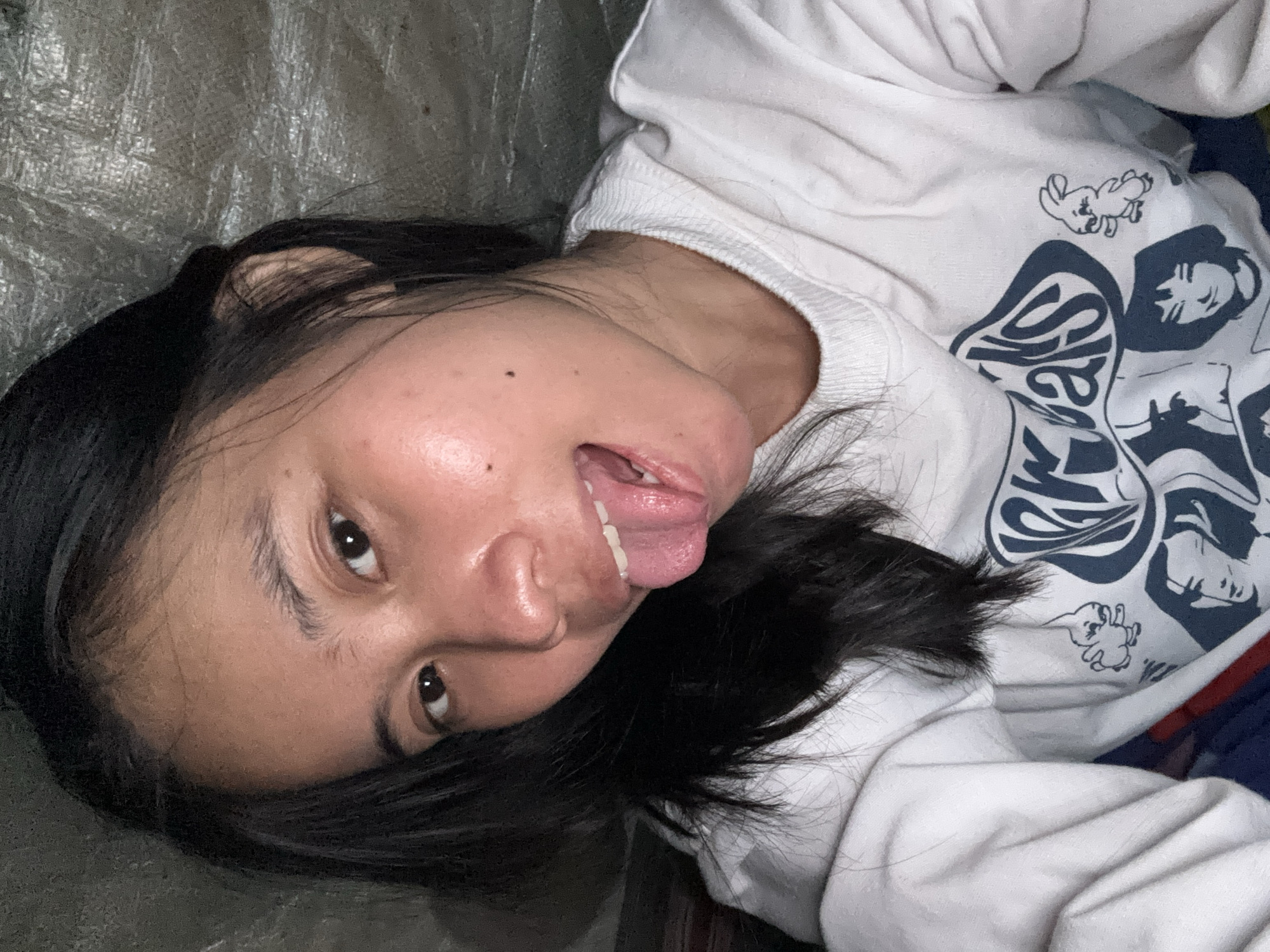 A young woman with black hair and brown eyes is shown from the chest up, sticking her tongue out at the camera. She is wearing a white sweatshirt with blue graphics. The background appears to be a textured, dark material. The image was taken indoors, with soft lighting. This scene is located in East Jakarta, Indonesia.