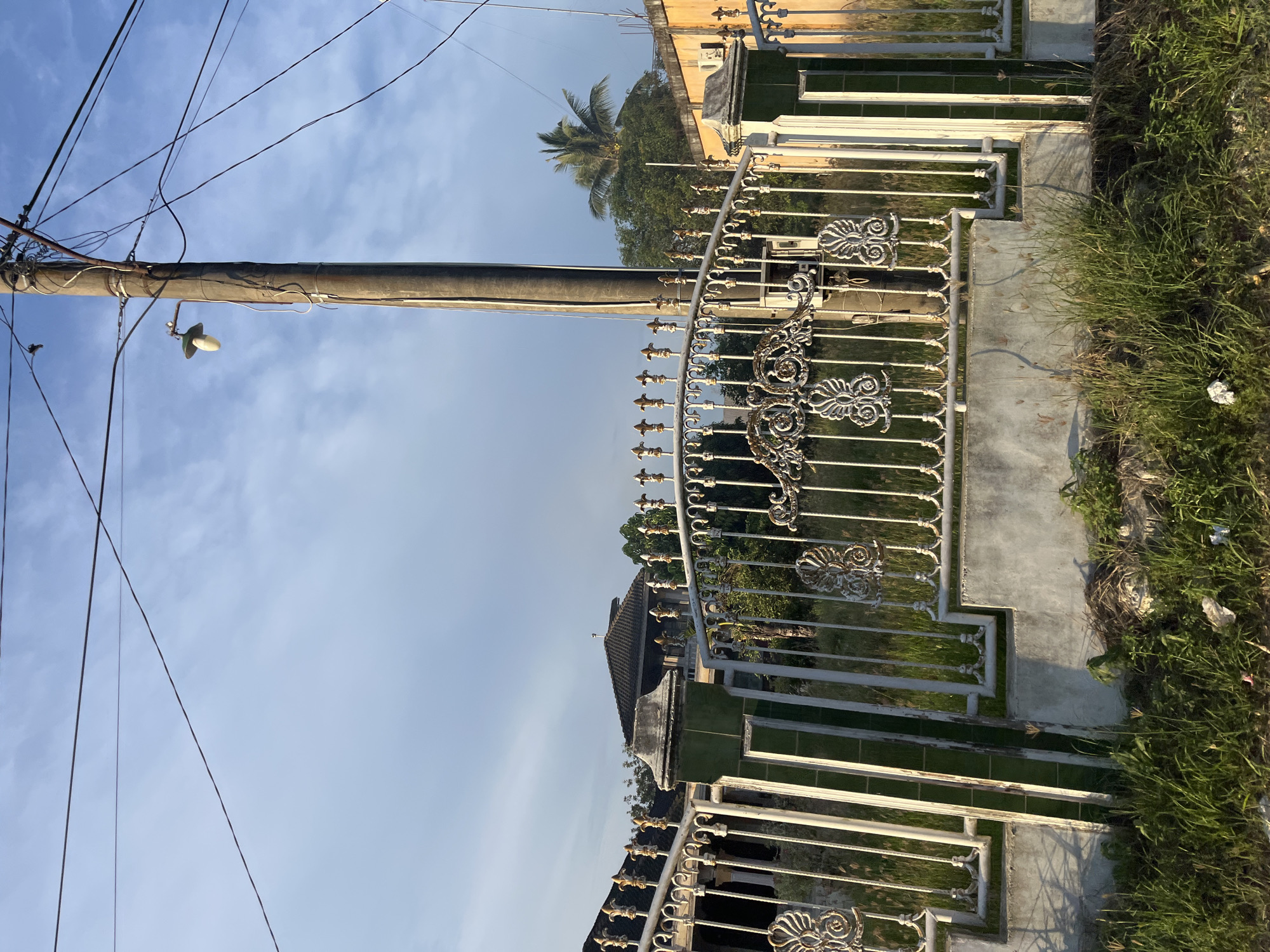 The image depicts a white metal fence with ornate scrolling designs and finials, set against a backdrop of a partly cloudy blue sky. A concrete base supports the fence, with overgrown grass and some litter at its foot. To the right of the fence, a weathered wooden utility pole stands, adorned with a street lamp and numerous electrical wires crisscrossing the frame. Behind the fence, a glimpse of a residential area is visible, featuring a building with a dark tiled roof and trees, including a palm tree. The scene is bathed in the warm light of what appears to be late afternoon or early morning sun. The location is identified as Seunebobaru, Indonesia.