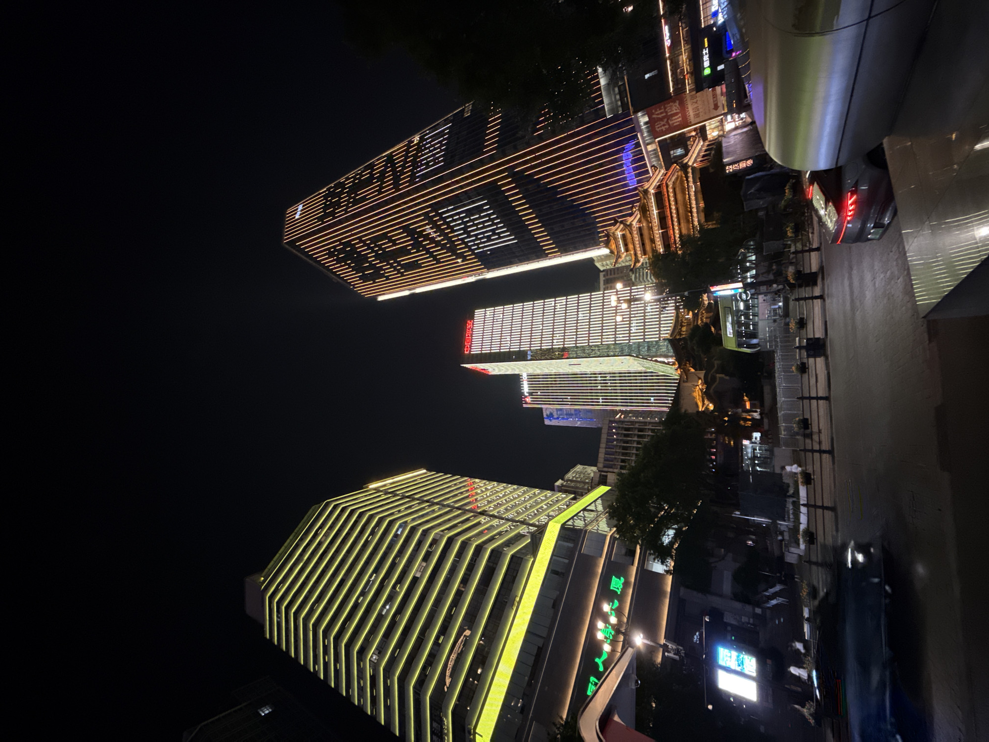 The image depicts a nighttime cityscape in Yuzhong District, China, characterized by illuminated skyscrapers and traditional architecture. In the foreground, a dark, blurred car moves from left to right, its brake lights illuminated. To the right, a large, curved, metallic structure partially obstructs the view. The midground features a street with a few blurred figures walking on the sidewalk and trees. Several multi-story buildings are prominent. The building on the far left is a tall skyscraper with horizontal yellow lighting. Adjacent to it, to the right, are two other skyscrapers: one with vertical orange and white lights, displaying text, and another to its left with similar vertical lighting and red Chinese characters. In front of these skyscrapers, a traditional multi-tiered, pagoda-style building with illuminated eaves is visible, surrounded by trees. The background is dominated by the dark night sky and the illuminated facades of the skyscrapers.
