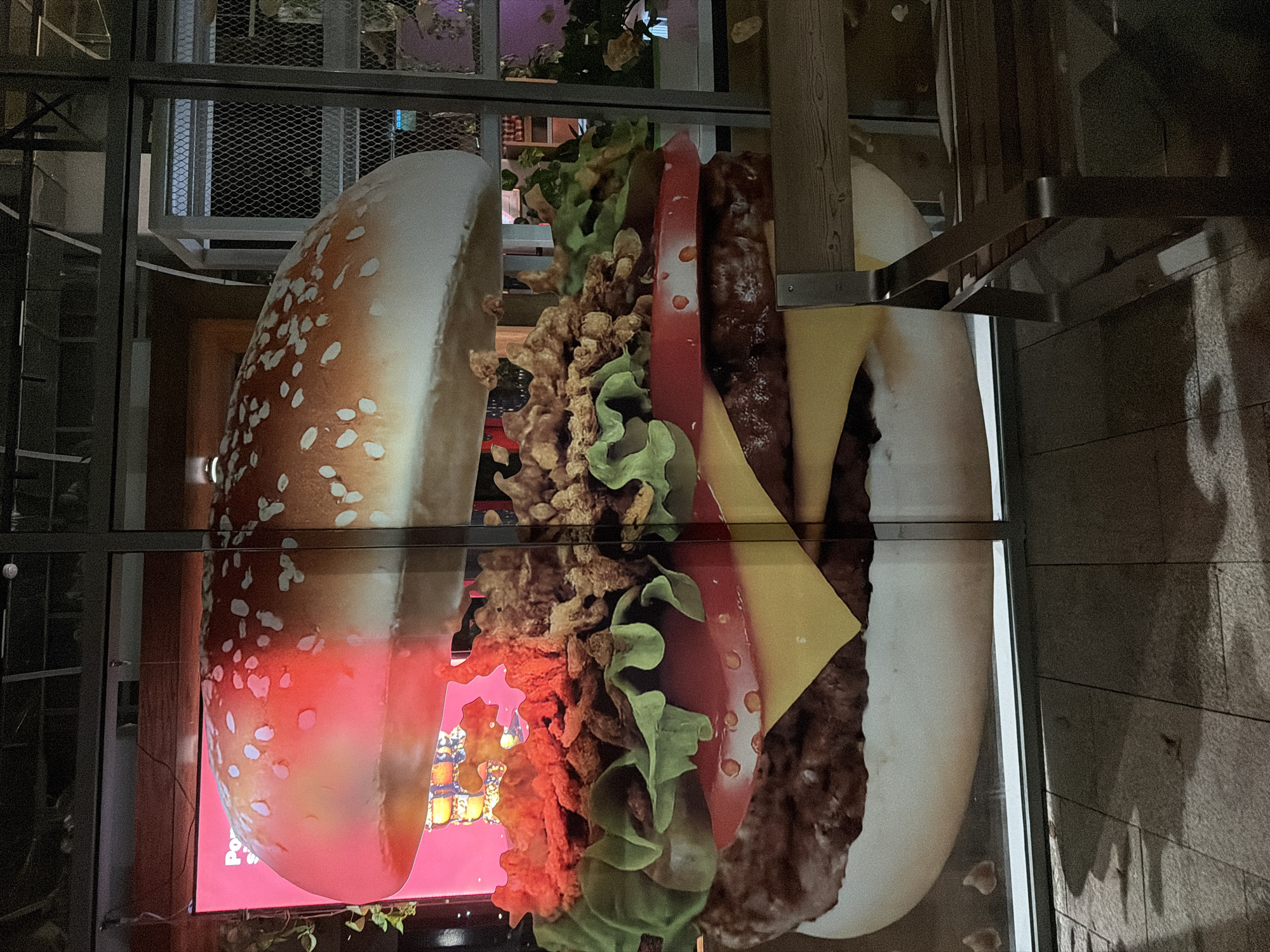 A large, photorealistic mural of a hamburger is displayed on a glass facade, possibly a storefront or window. The hamburger is depicted with a sesame seed bun, lettuce, tomato, cheese, and fried onions. The mural appears to be illuminated by red light from behind, giving the top bun a warm glow. In the foreground, a portion of a wooden bench with a metal frame is visible. The background through the glass shows a dimly lit interior with metal structures and greenery. The ground in front of the facade is paved with stone tiles and shows shadows. This scene is located in Chinarkent, Uzbekistan.