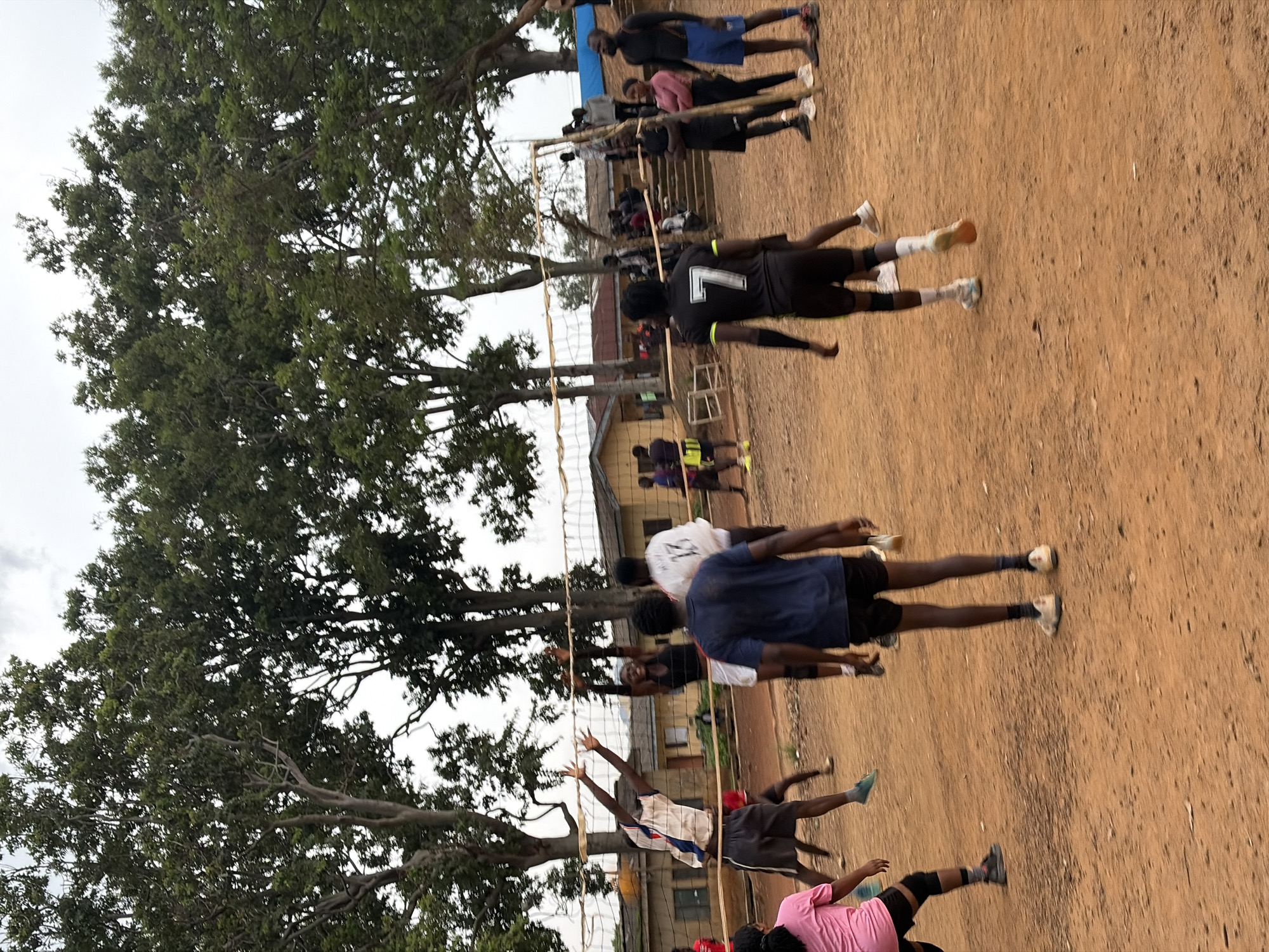 In Rafin Par, Nigeria, on a dusty, outdoor volleyball court surrounded by trees and buildings, a volleyball game is in progress. Several young men and a woman are actively engaged in the game. One man in a white and blue jersey and dark shorts is jumping to hit the ball, while another in a black sleeveless shirt and white shorts is also reaching up. A man in a dark blue shirt and dark shorts stands facing away from the camera, and another in a black jersey with the number 7 on the back is also visible. A woman in a pink shirt and black shorts is on the left side of the court. Other players and spectators are scattered around the court and in the background. The court surface is dirt, and a volleyball net is strung across the middle. The sky is overcast.