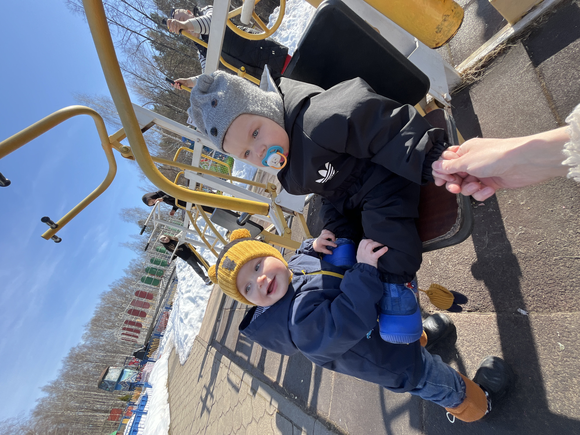 Two young children are sitting on a piece of playground equipment. The child closer to the viewer is wearing a blue jacket, blue shoes, and a yellow knit hat with pom-poms. The child is smiling and looking directly at the camera. The other child is wearing a black jacket, black pants, and a gray knit hat with ear-like protrusions. The child has a pacifier in their mouth and is looking to the side. An adult hand is holding the sleeve of the child in the black jacket. The playground equipment appears to be a seesaw-type ride with yellow and white metal supports. In the background, there is a playground with various colorful structures, trees, and patches of snow. Based on other available information, it is likely the location is in the city of Chaikovsky, Russia.