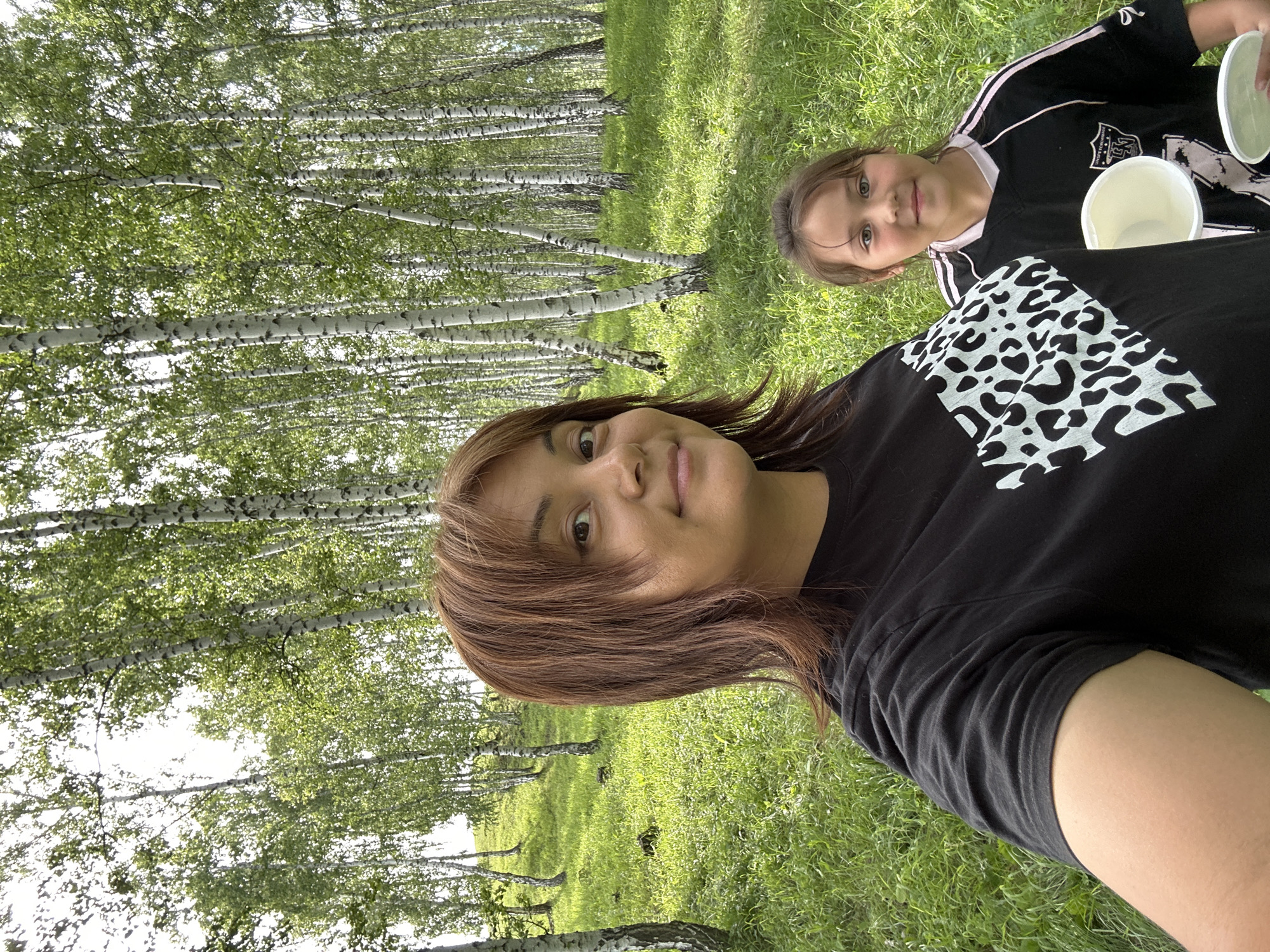 A woman and a young girl are standing in a birch forest. The woman is in the foreground, facing the camera and smiling. She has medium-length brown hair and is wearing a black t-shirt with a white leopard print design. The girl is behind the woman, also facing the camera and smiling. She has brown hair and is wearing a black shirt with pink accents. She is holding two white bowls. The forest is dense with tall, slender birch trees with white bark and green leaves. The ground is covered in lush green grass. The lighting suggests it is daytime.