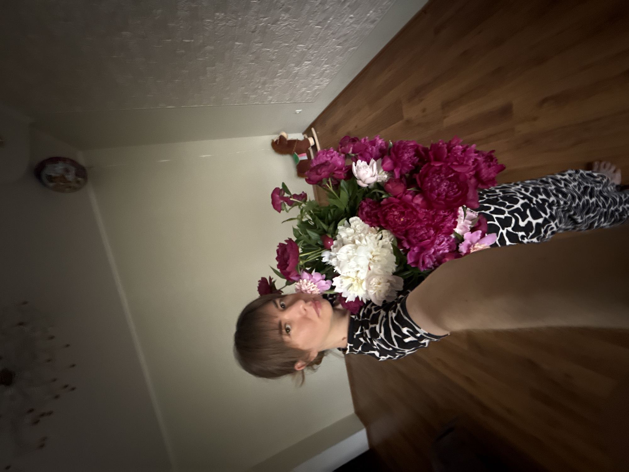 A woman wearing a black and white patterned top is holding a large bouquet of pink and white peonies. She is positioned indoors, in front of a wall, adjacent to a wooden floor. A stuffed animal is on the wall, and a small decorative plate is above it. The location is Unknown, Unknown.