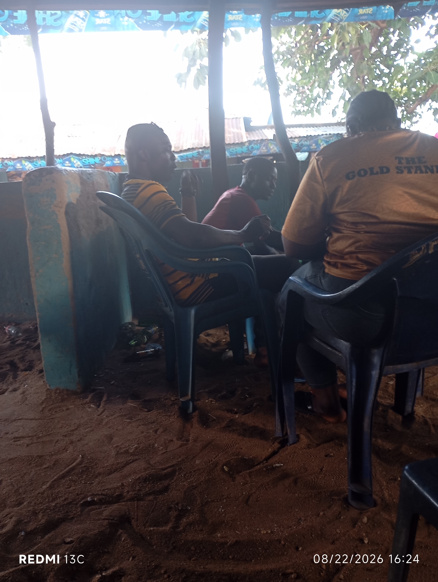 Three individuals are seated in chairs outdoors, likely in a rural or less developed setting, with sandy ground. The person on the left, wearing a striped yellow and black shirt, is facing left. The person in the middle, wearing a red shirt, is partially visible and appears to be looking downwards. The person on the right, wearing a yellow shirt with "THE GOLD STAND" printed on the back, is facing away from the camera. The environment suggests a casual gathering or rest stop, possibly at a roadside establishment, with some debris on the ground. The background includes a corrugated metal roof and vegetation, indicating an outdoor, possibly tropical or subtropical, location. The presence of the date and time stamp "08/22/2026 16:24" and "REDMI 13C" suggests this image was taken with a mobile phone. The image does not provide specific visual cues to confirm the location as Shendam, Nigeria, but the general context could be consistent with such a setting.